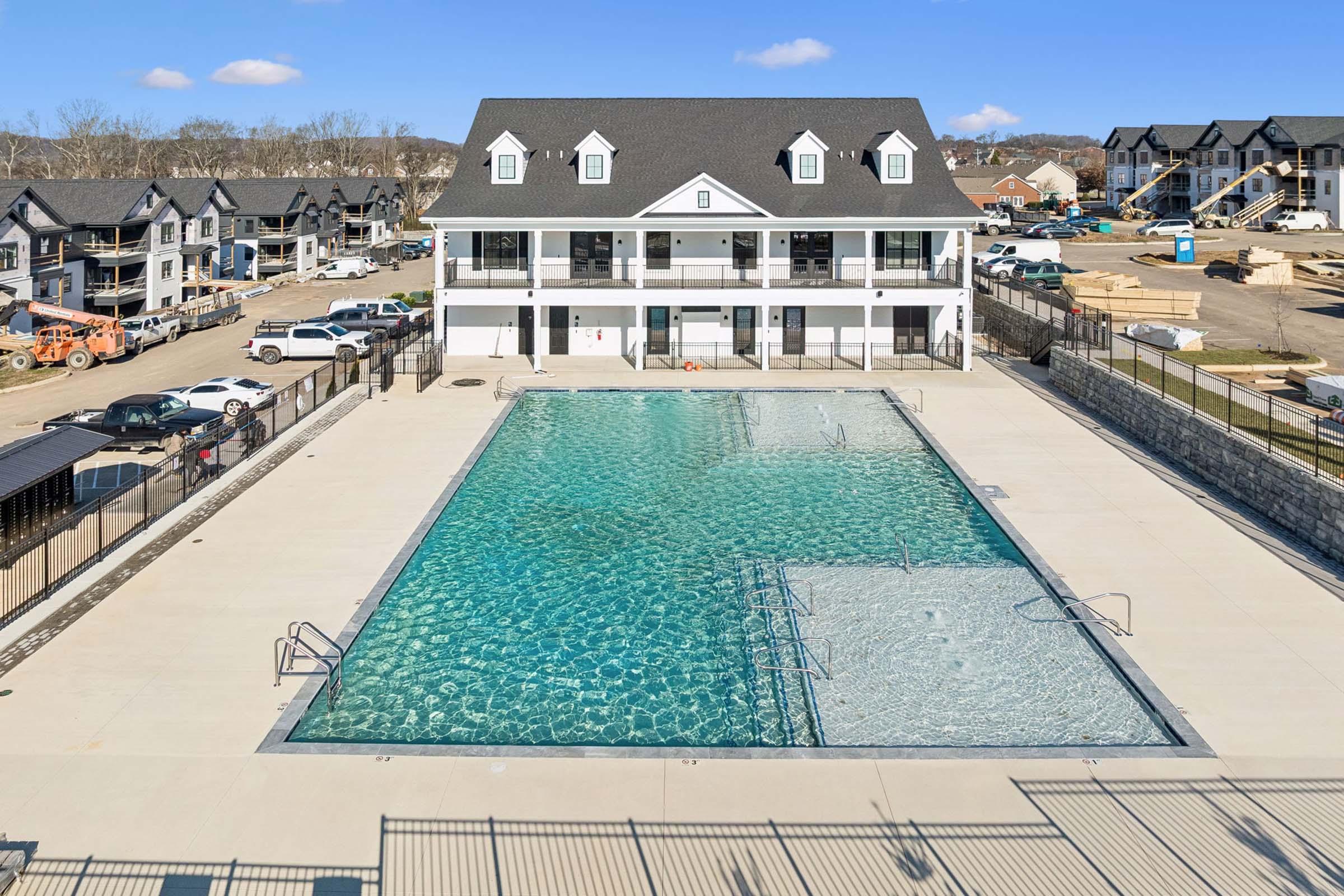 A large swimming pool with clear blue water in front of a modern two-story building. The pool features a shallow area and surrounded by a fenced patio. In the background, there are several buildings under construction and parked vehicles, with a clear sky above.