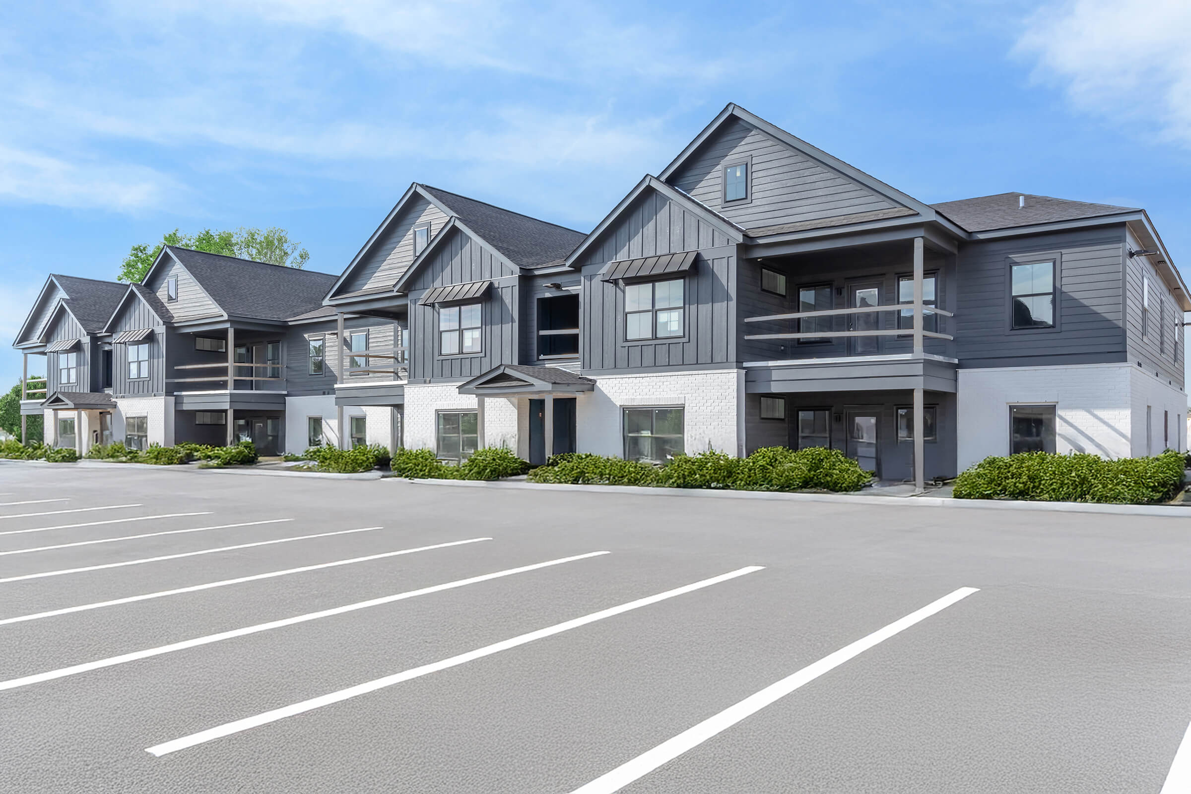 A modern multi-unit residential building with gray and white exterior. The structure features balconies and large windows. A spacious parking area with marked spaces is in the foreground, set against a clear blue sky and greenery nearby.