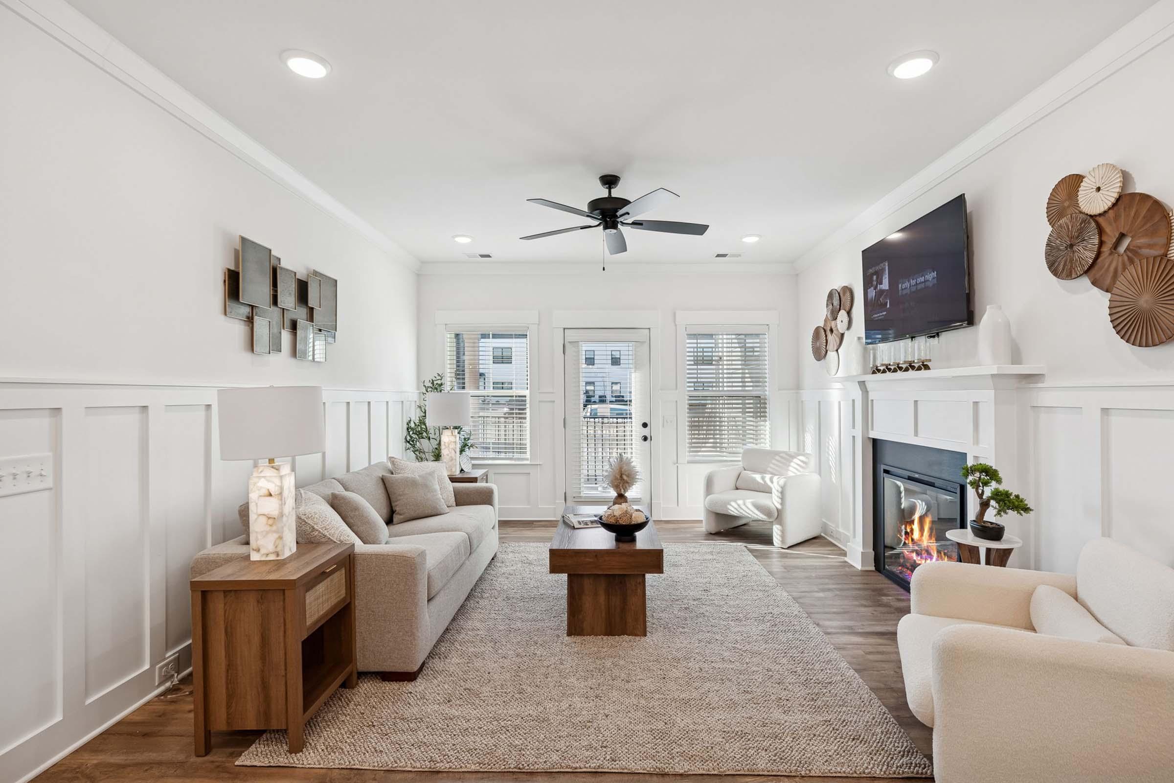 A modern living room with white walls and sleek design. Features include a cozy seating area with light-colored sofas, a wooden coffee table, and decorative wall art. A ceiling fan hangs above, and a flat-screen TV is mounted on the wall. Large windows allow natural light in, enhancing the warm atmosphere.