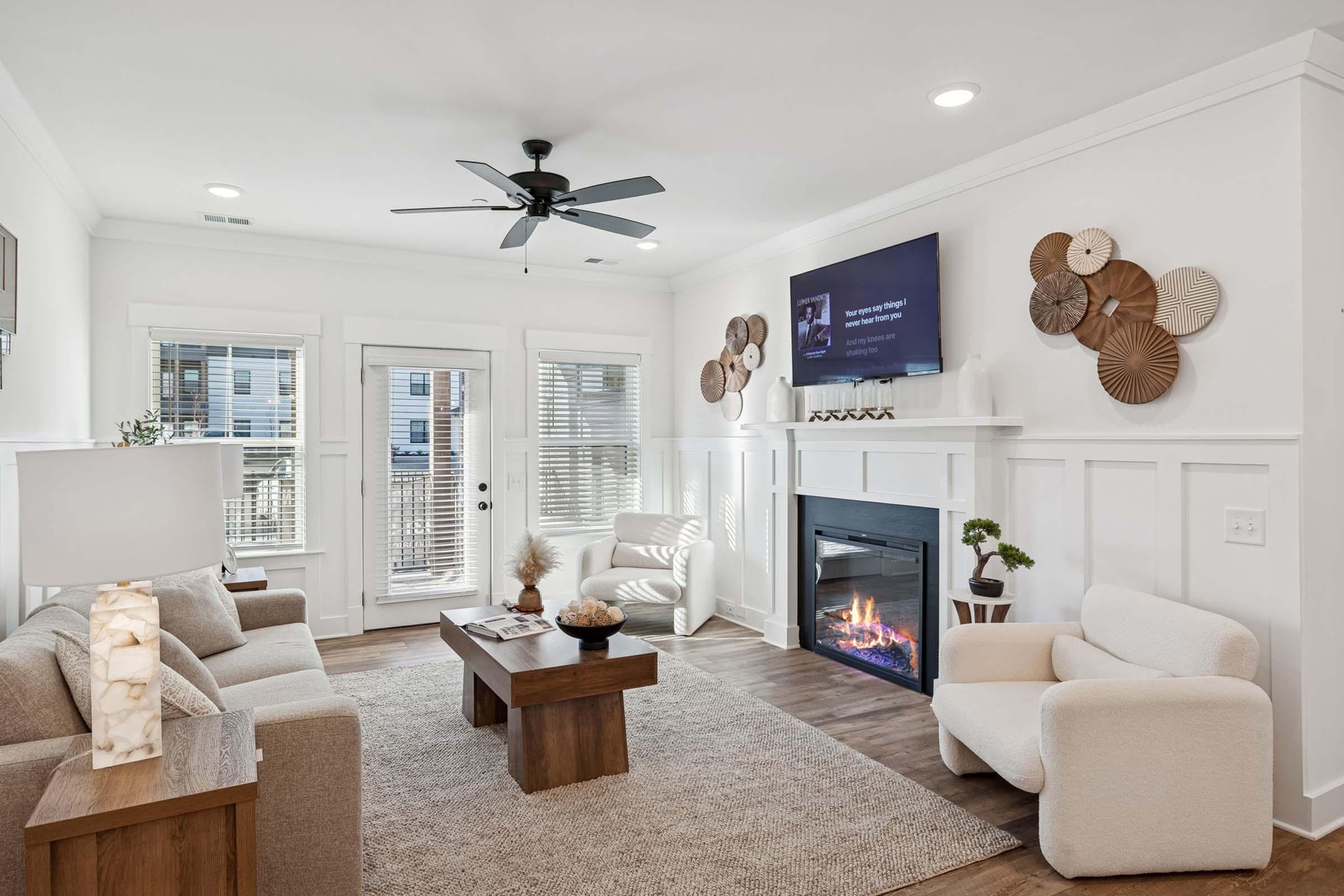 A modern living room featuring a cozy seating arrangement with a sofa, armchair, and coffee table. A fireplace sets a warm ambiance, and a television is mounted on the wall. Natural light flows through the windows, and decorative wall art adds a touch of style.