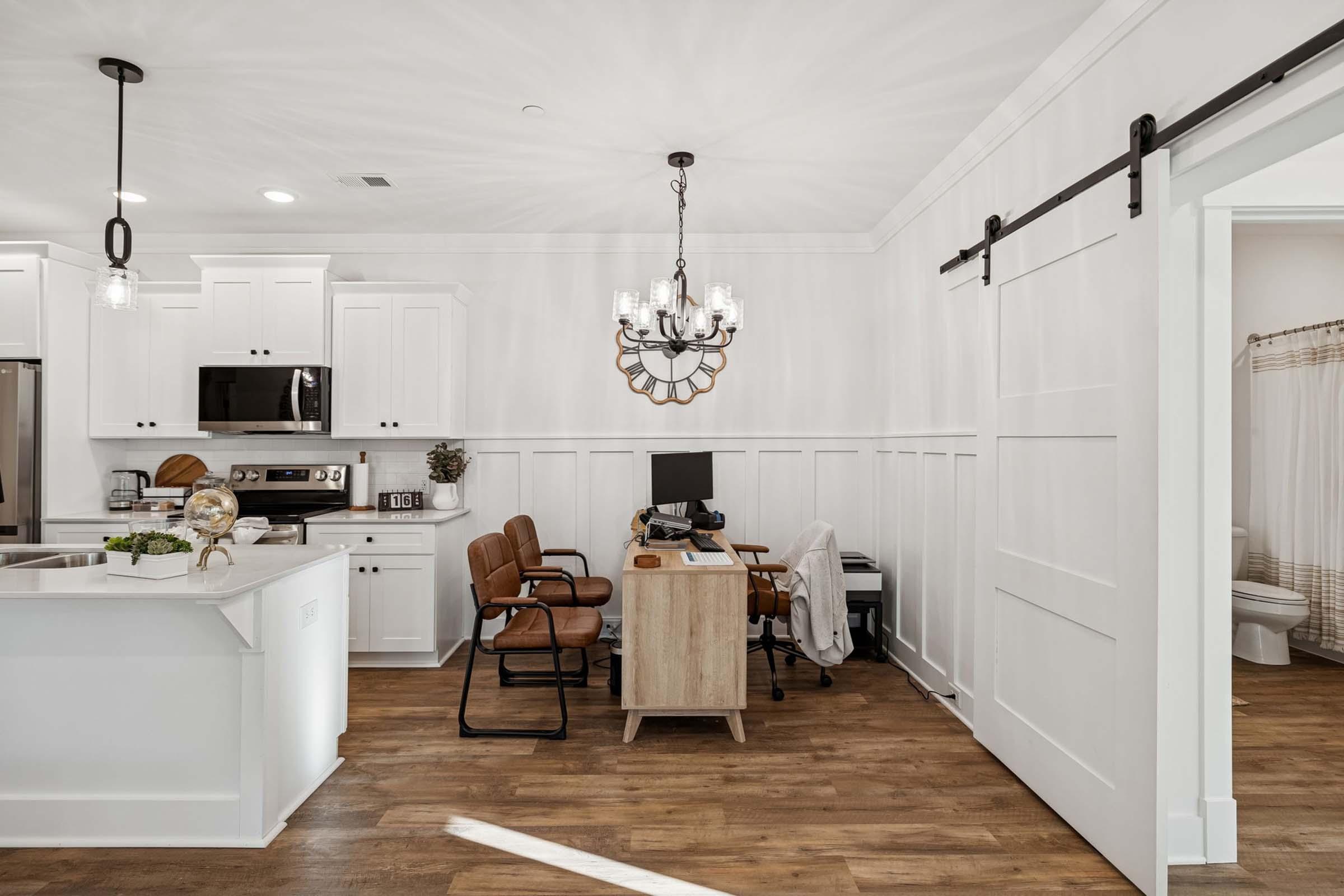 A modern kitchen and home office space featuring white cabinets, a wooden dining table, and a stylish chandelier. The workspace includes a desk with a computer and comfortable chairs, along with a sliding barn door and a glimpse of a bathroom in the background. Natural light illuminates the room.