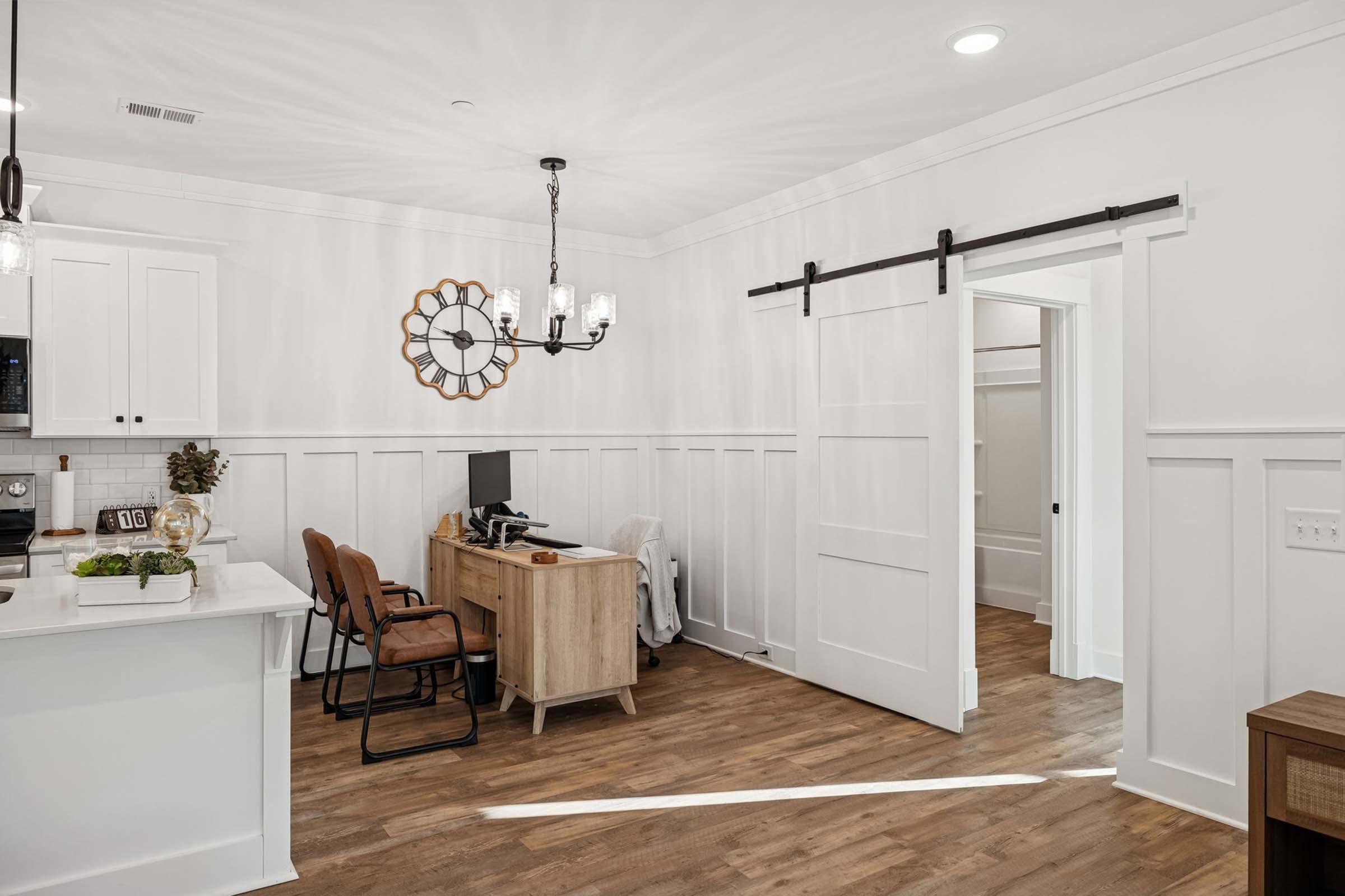 A modern office space featuring a wooden desk with chairs, a large wall clock, and white wainscoting. The room has a sliding door leading to a hallway, and the floor is made of light-colored wood. Natural light is coming in, creating a bright and inviting atmosphere.