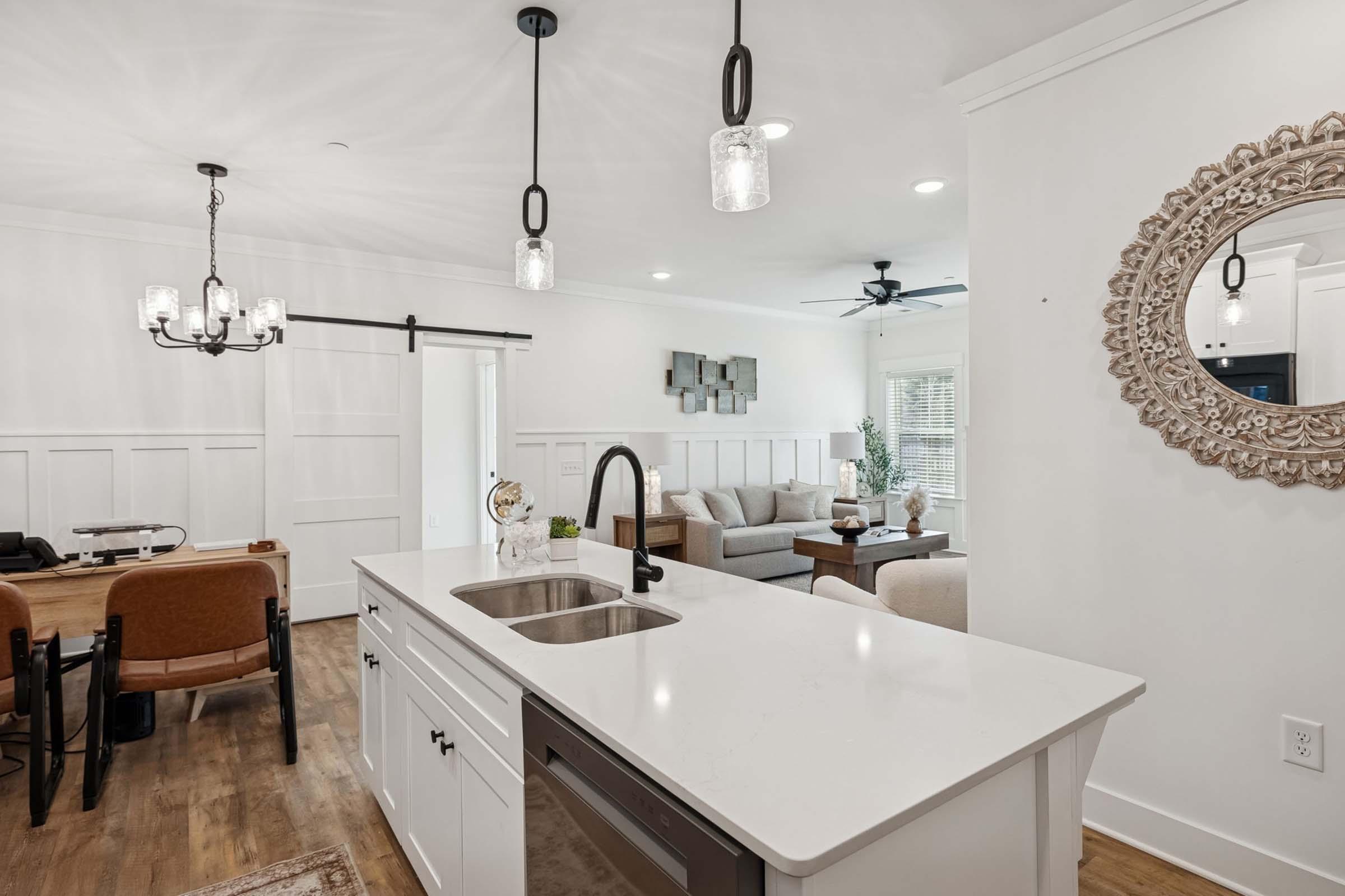 A modern, bright interior featuring an open kitchen with a white countertop and sink. The dining area has a wooden table with chairs. A cozy living room with a light-colored sofa and decorative pillows is visible in the background. Stylish light fixtures and a round mirror enhance the contemporary decor.