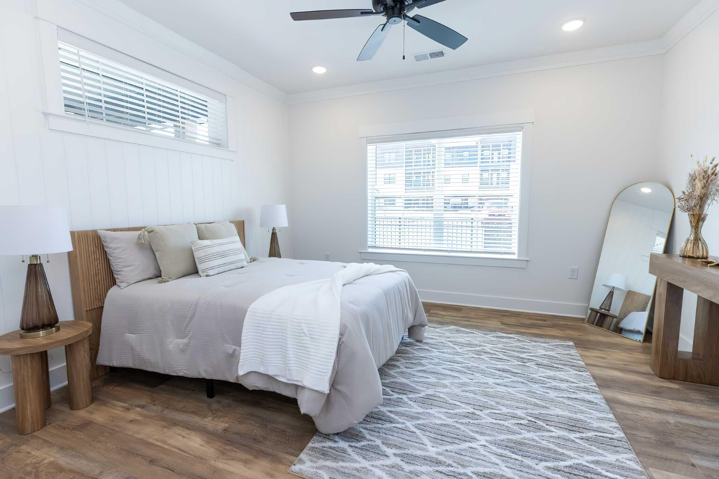 A modern bedroom featuring a neatly made bed with light-colored linens, two bedside tables with lamps, and a large mirror on a console table. The room has natural light coming through window blinds and light hardwood flooring, creating an airy and inviting atmosphere.
