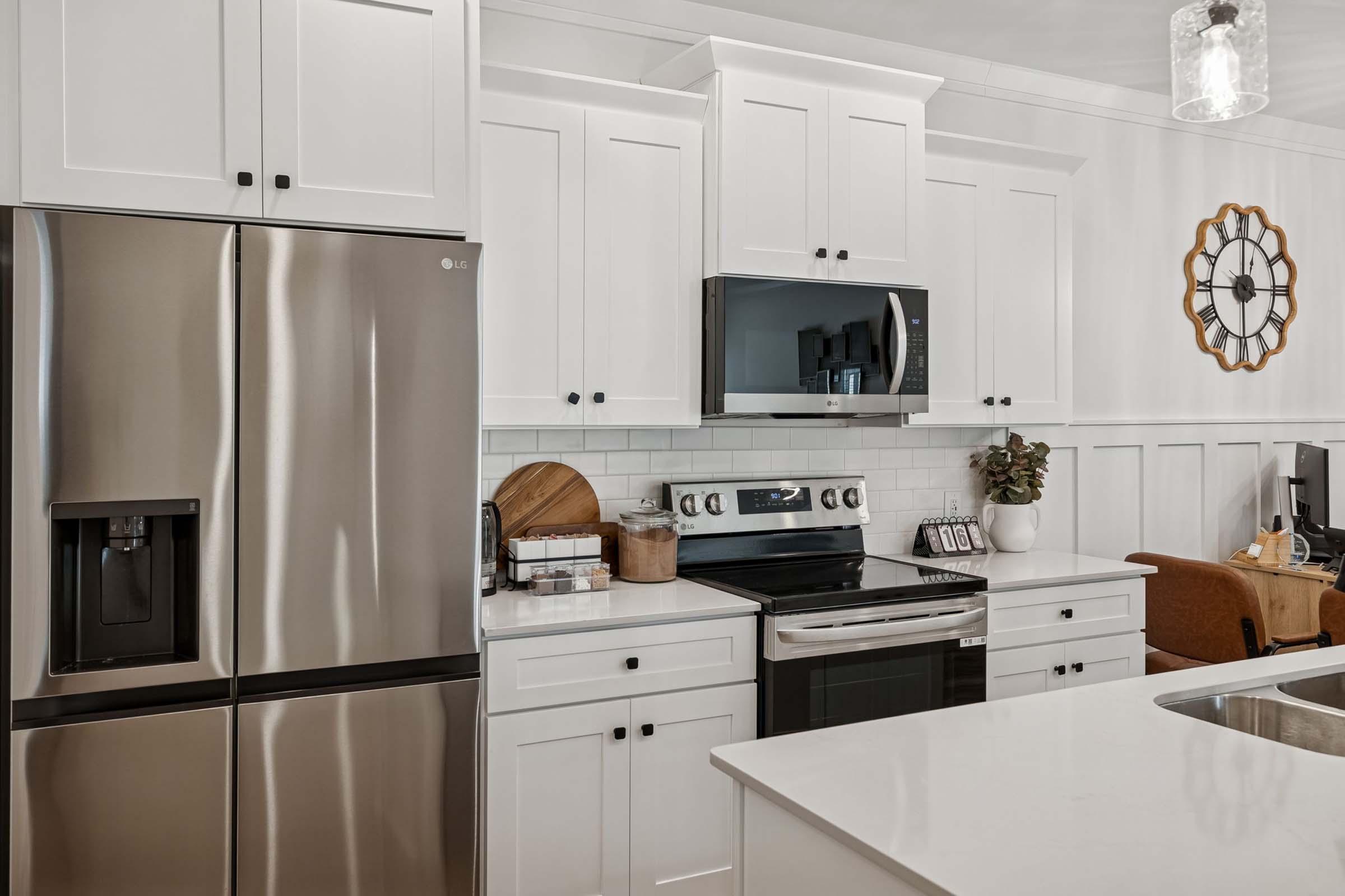 Modern kitchen featuring stainless steel appliances, including a refrigerator and oven, with white cabinetry and a composite sink. A black microwave is mounted above the stove, and there's a decorative clock on the wall. The space includes a small dining area with a chair and a plant on the countertop.
