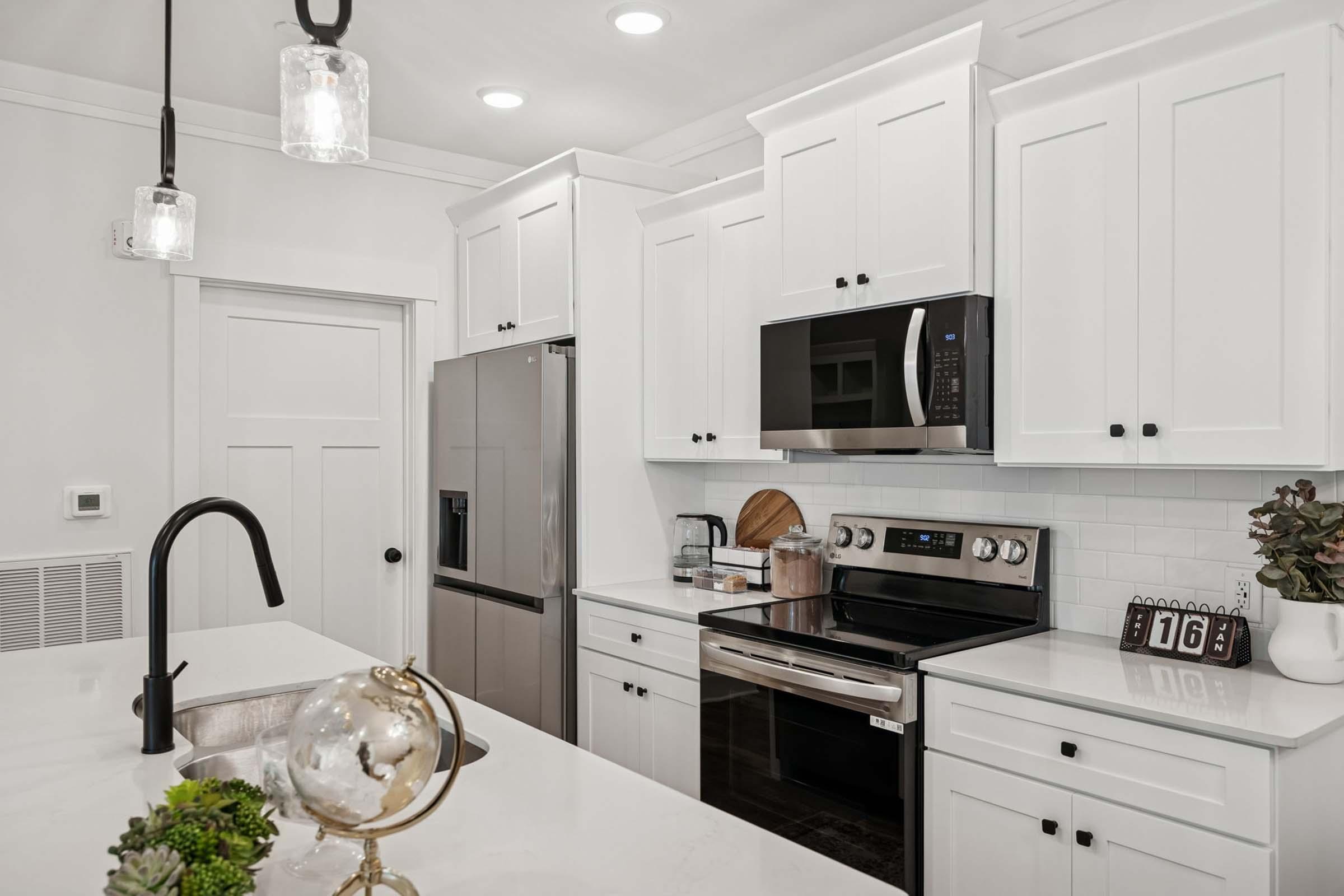 A modern kitchen featuring white cabinets, stainless steel appliances, and a sleek countertop. The space includes a sink, a refrigerator, and an oven. Decor elements include a small globe and plants, with pendant lighting and a wall clock visible. The overall design is bright and contemporary.