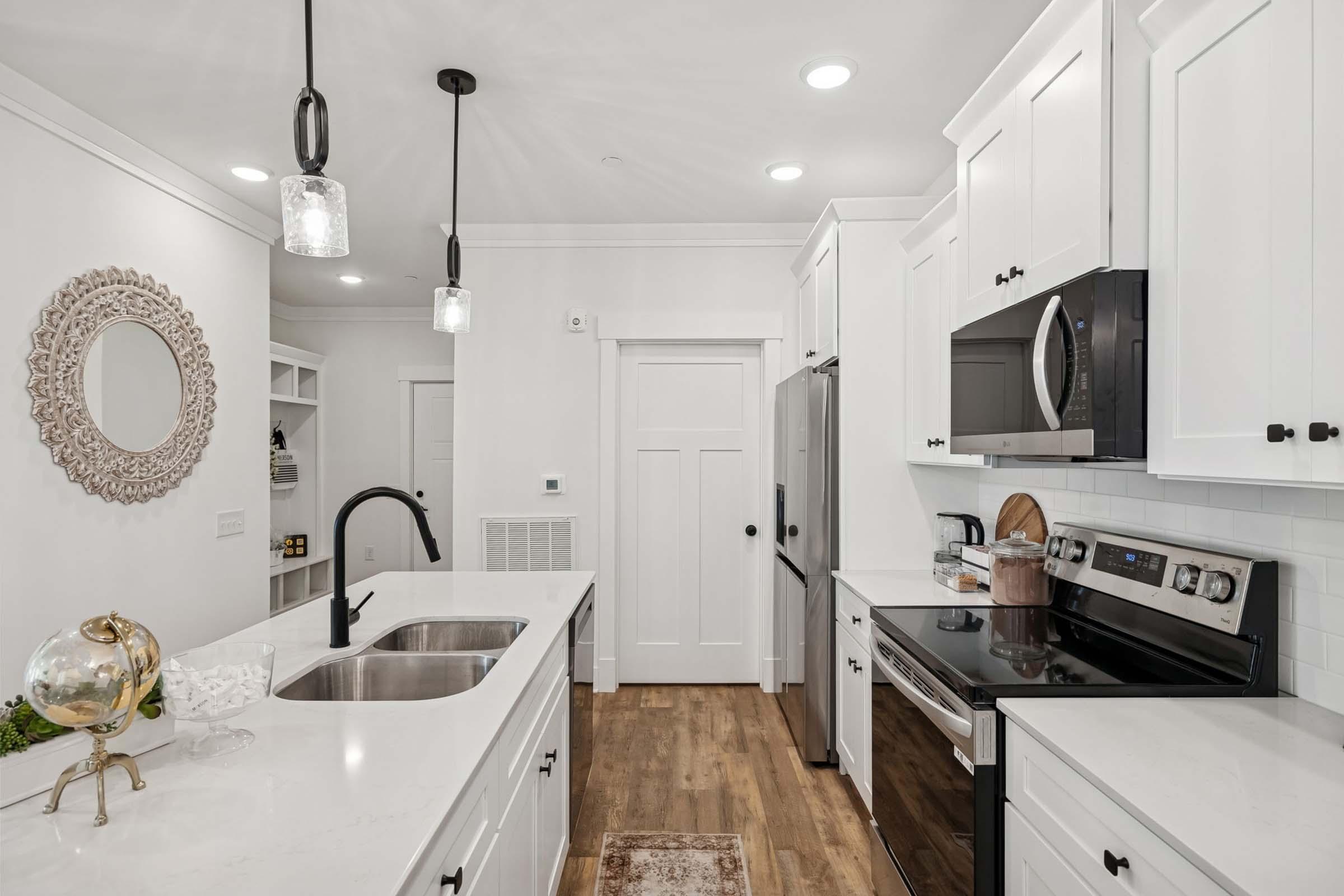 A modern kitchen featuring white cabinets, a stainless steel stove, and a refrigerator. The island sink has a sleek black faucet. Pendant lights hang above, illuminating the space. A round decorative mirror and shelves are visible in the background. The floor is made of warm wood tones, adding coziness to the contemporary design.