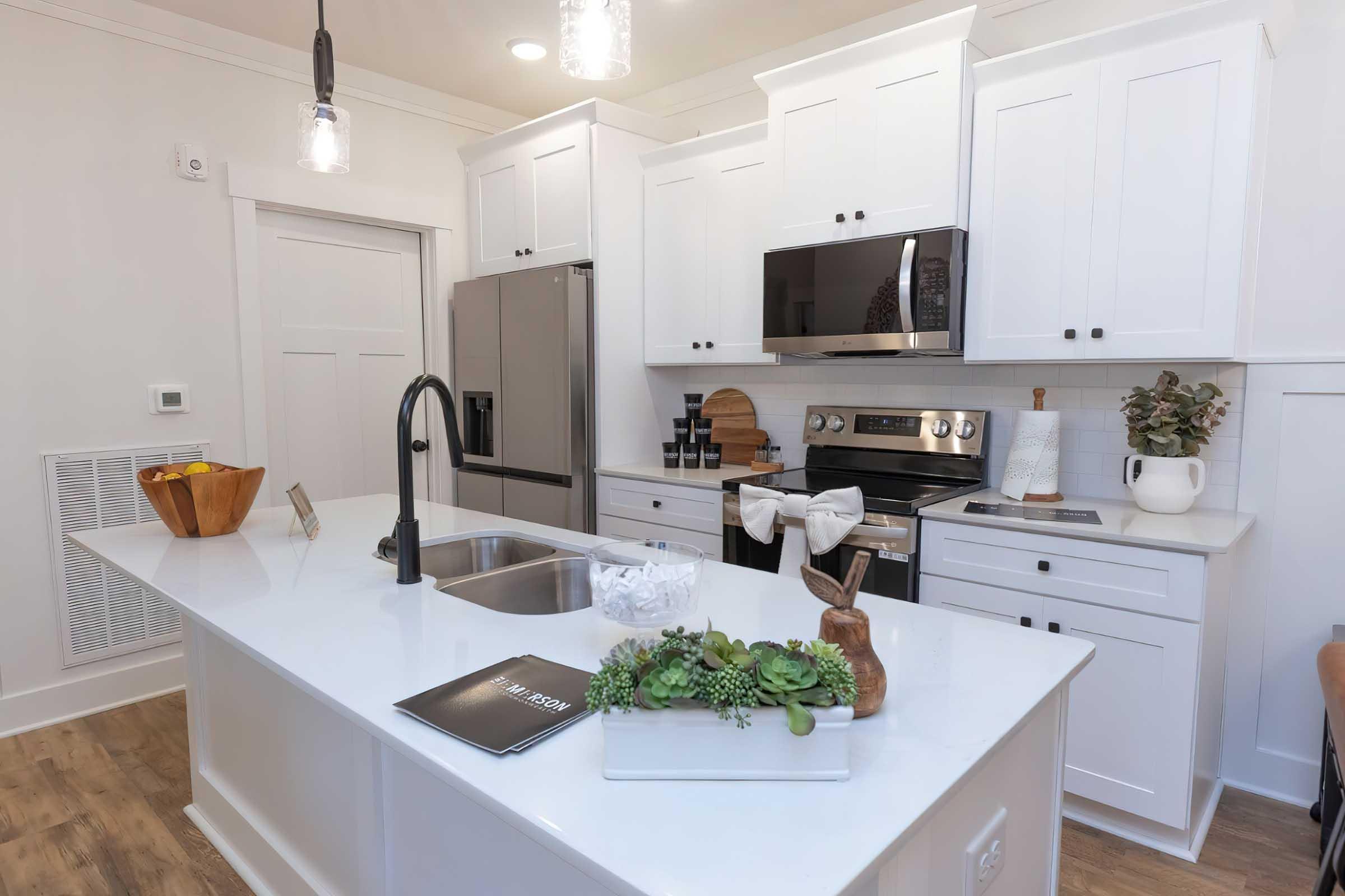 Modern kitchen featuring white cabinets, stainless steel appliances, and a center island with a sleek countertop. Decor includes a bowl of wooden spoons, a bunny figurine, succulents, and a notepad. Bright lighting enhances the spacious and contemporary design.