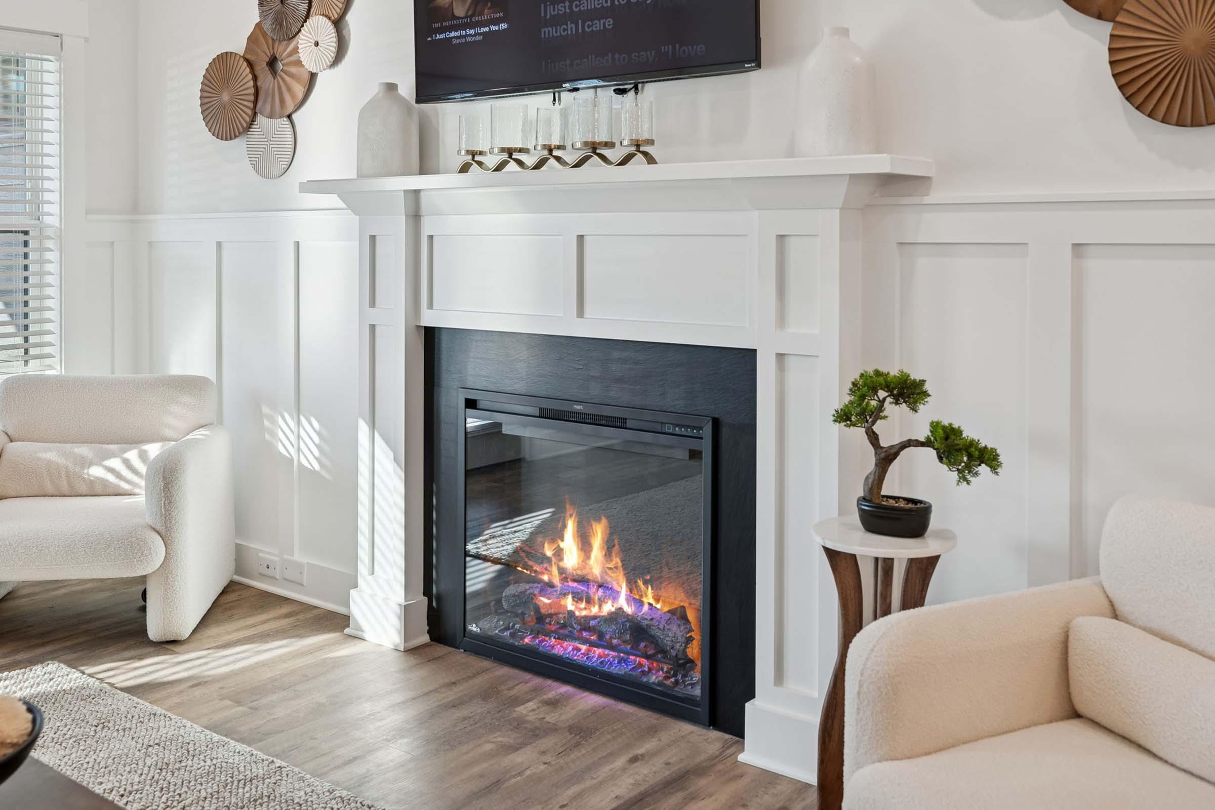 A cozy living room featuring a modern electric fireplace with a glowing flame, a white mantel, and decorative wall art. Soft furnishings include two light-colored chairs and a small side table with a potted plant. Natural light filters through nearby windows, enhancing the inviting atmosphere.