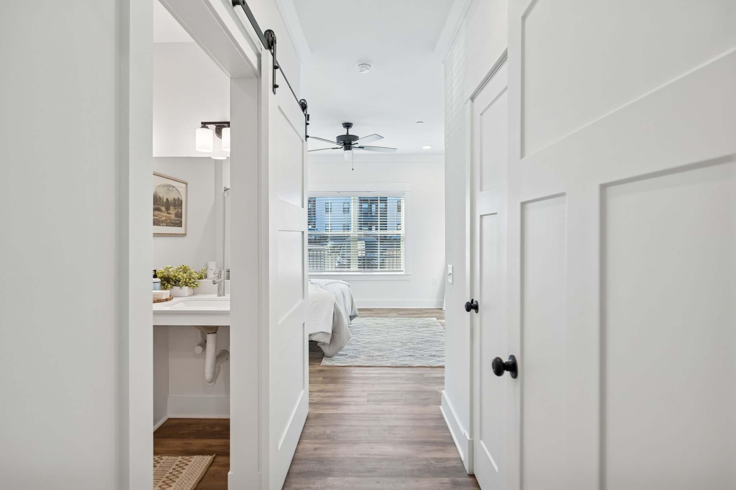 A well-lit hallway featuring white sliding doors leading to a bedroom. On one side, a bathroom area is partially visible with a sink and decor. The opposite side shows a cozy bedroom with a light color scheme and a window letting in natural light. The floor is wooden, enhancing the inviting atmosphere.