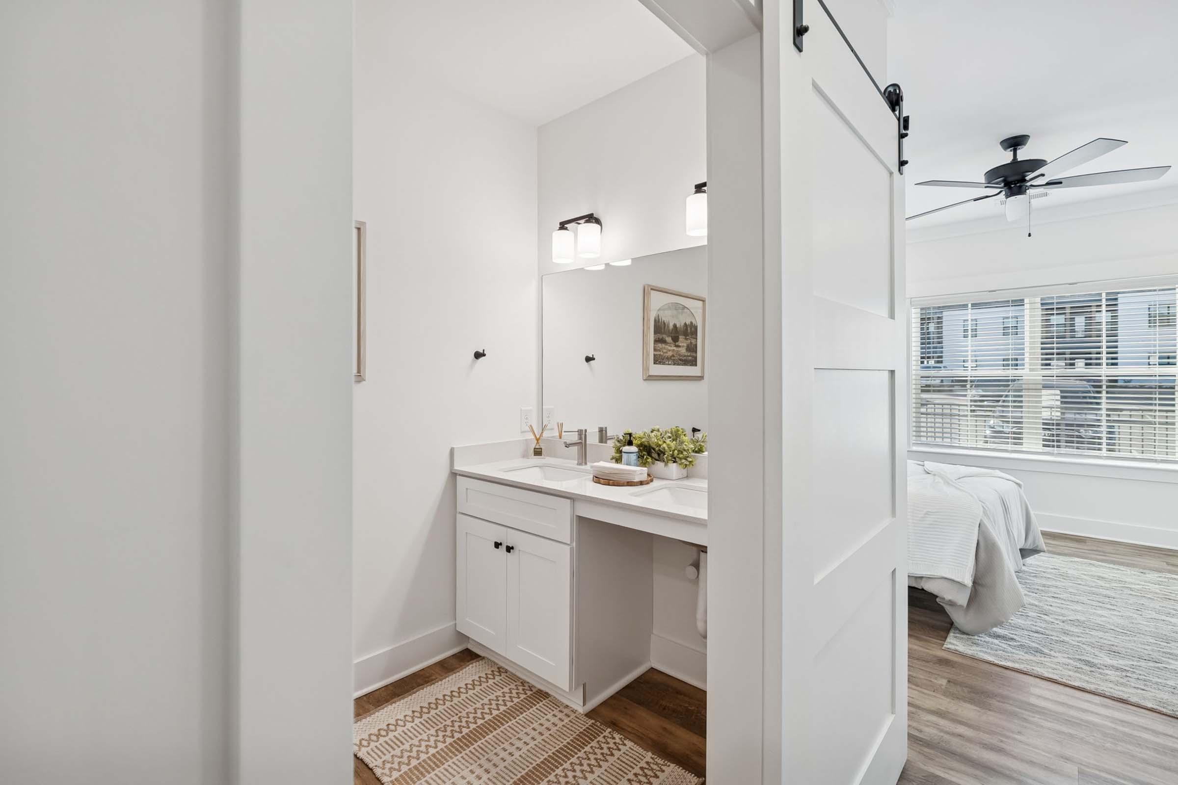 A modern bathroom featuring a white vanity with a sink, framed artwork on the wall, and decorative items. The space is well-lit with two ceiling lights and a ceiling fan. A small window allows natural light, and the adjacent room visible in the background has a bed with light-colored bedding.