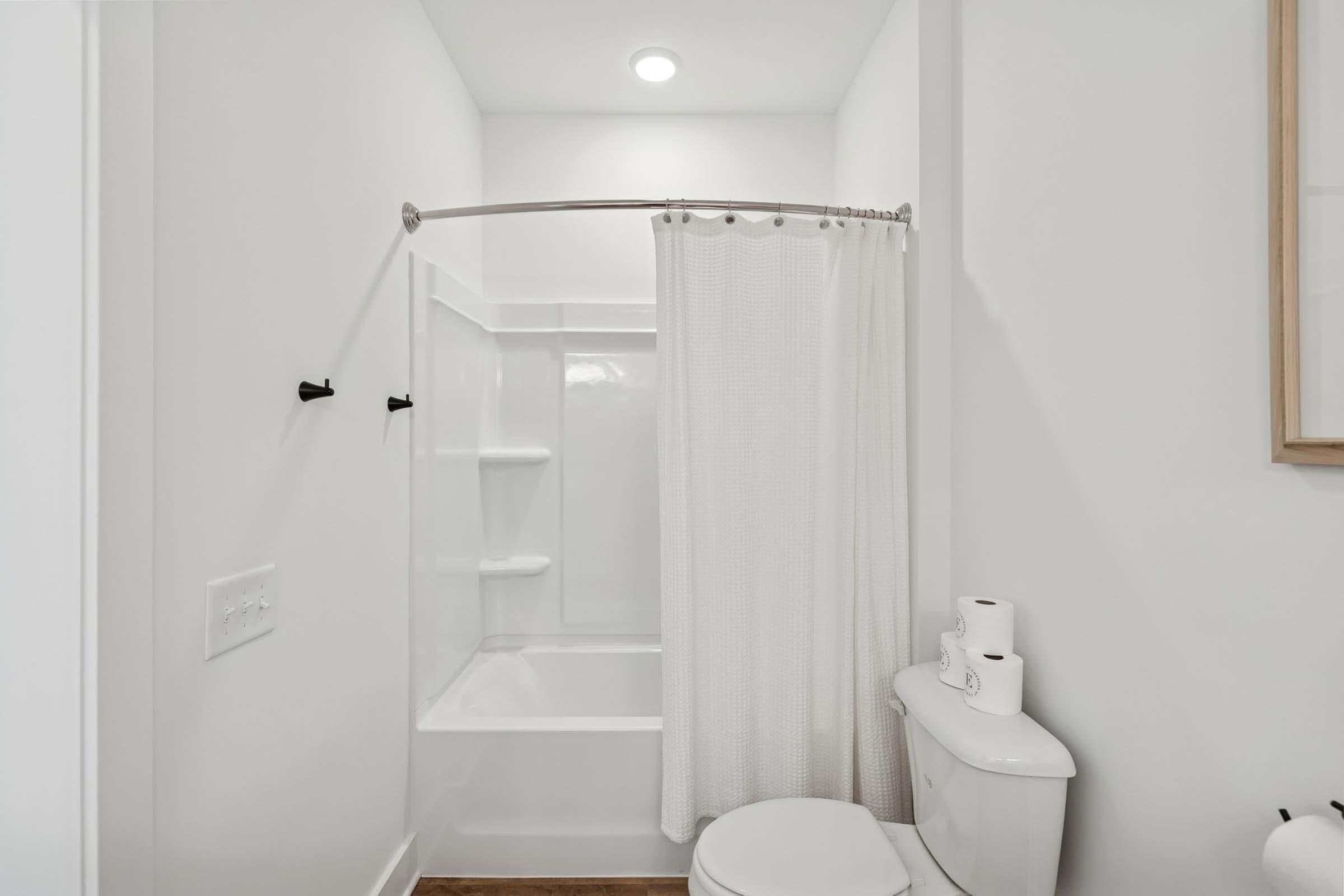 A clean and modern bathroom featuring a white bathtub with a shower curtain, a toilet, and shelves. The walls are light-colored, and the space includes basic amenities like toilet paper. The floor is wooden, adding warmth to the overall aesthetic.