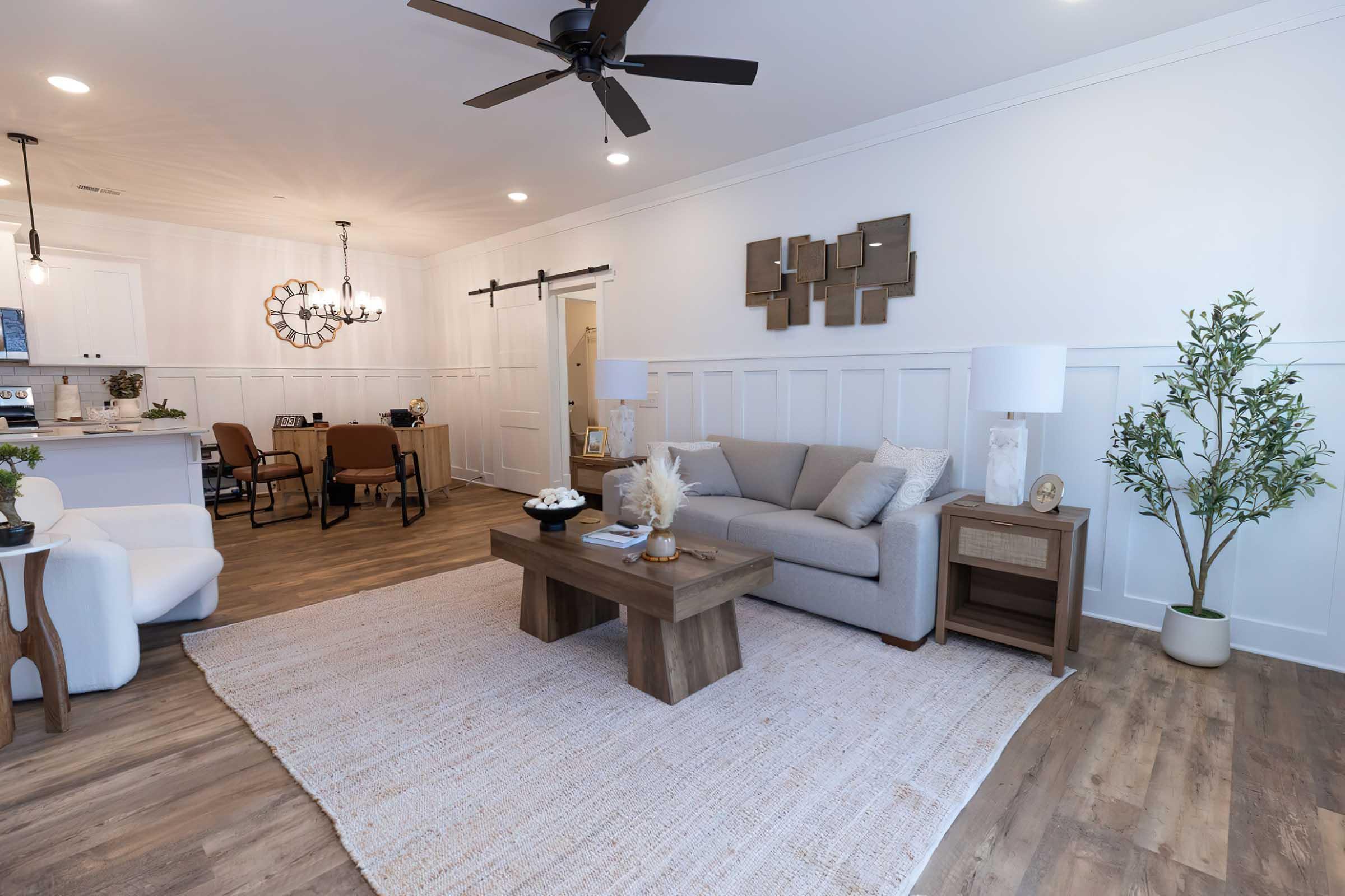 A modern living room featuring a light gray sofa, a wooden coffee table, and decorative plants. The space is illuminated with ceiling lights and has a textured area rug on the wooden floor. A dining area with brown chairs and a stylish light fixture is visible in the background, creating a cozy and inviting atmosphere.