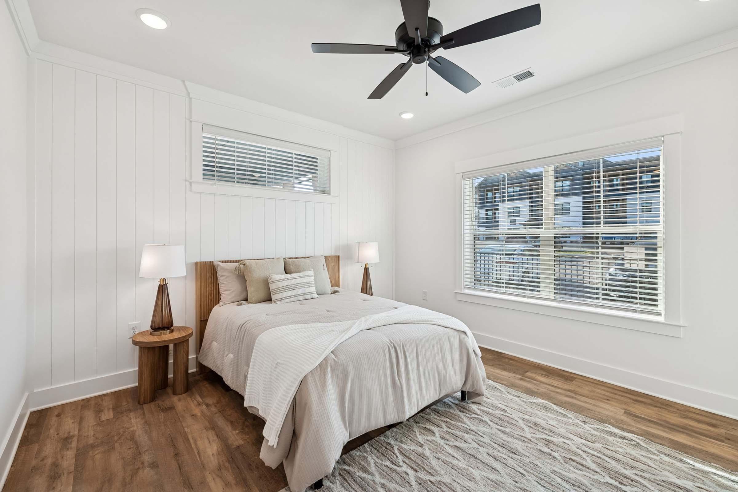 Cozy and modern bedroom featuring a wooden bed frame with neutral bedding, two bedside lamps on wooden tables, a ceiling fan, and large windows with white blinds. The room has light-colored walls and a soft area rug, creating a warm and inviting atmosphere.
