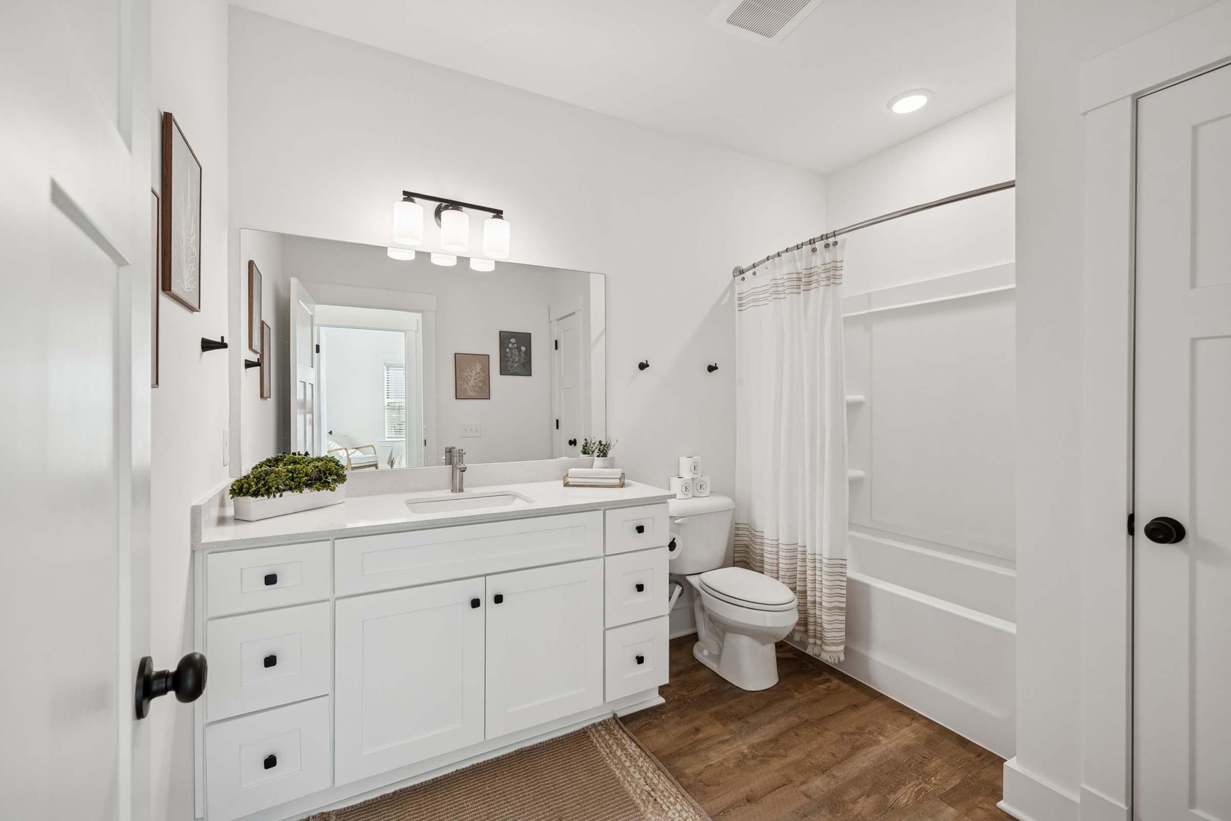 A modern bathroom featuring a white vanity with a large mirror, a sink, and drawer storage. There is a shower with a curtain, a toilet, and decorative framed art on the walls. The flooring is wooden, and there are bath essentials displayed, creating a clean and inviting space.