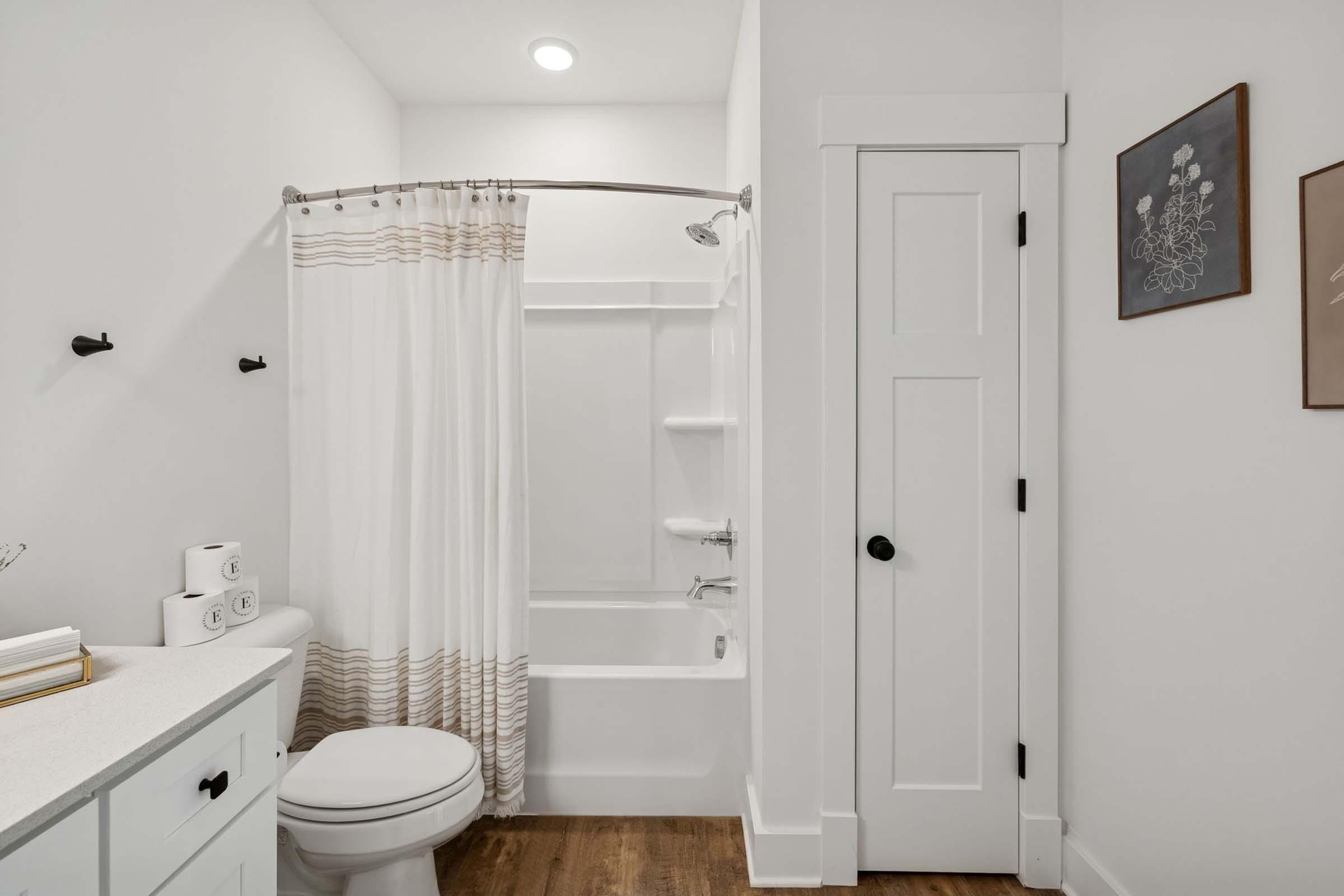 A clean, modern bathroom featuring a tub/shower combo with a white curtain, a toilet, and a small vanity with neatly arranged towels. The walls are painted white, and there's a door leading to another area. Art is displayed on the wall, adding a touch of decor. The flooring is made of wood.