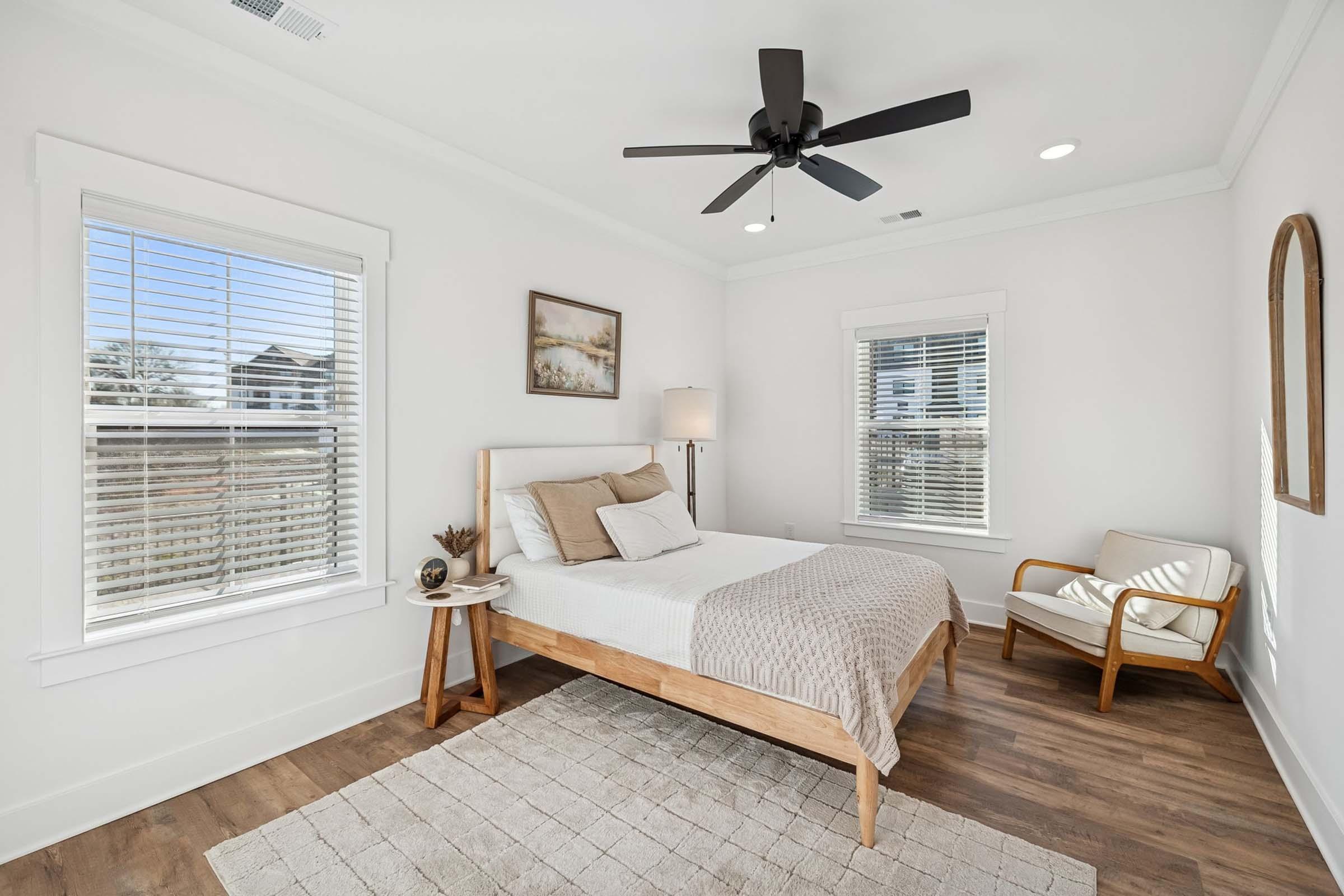A modern bedroom featuring a wooden bed with a light-colored duvet, a small side table with decorative items, and a cozy armchair. Large windows allow natural light to fill the space, highlighting the neutral color palette. A ceiling fan adds functionality to the airy atmosphere.