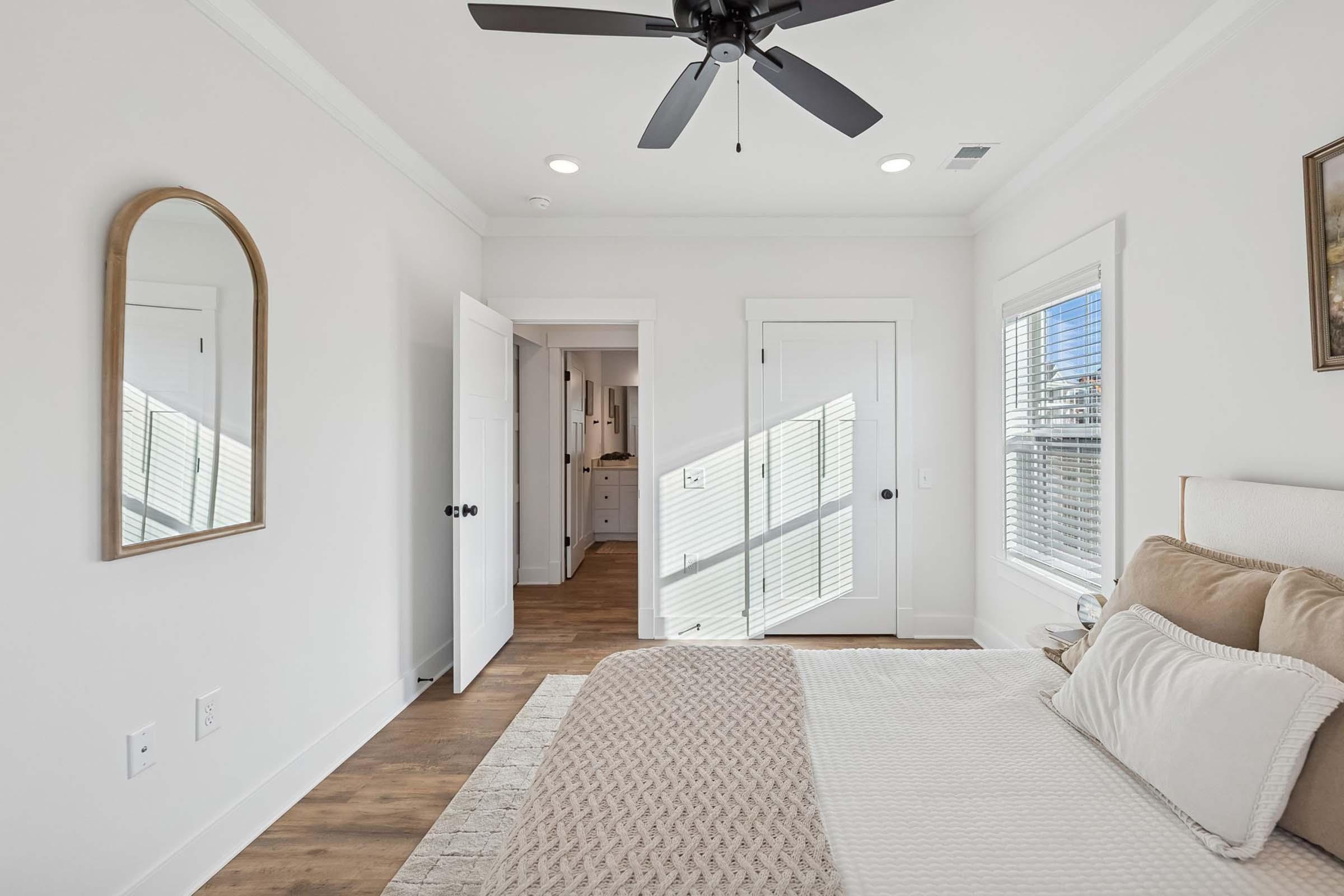A bright, modern bedroom featuring a beige bed with decorative pillows, a large mirror on the wall, and a ceiling fan. Light streams through windows, illuminating the room. A doorway leads to a hallway that connects to a bathroom, with light-colored walls and wooden flooring throughout.