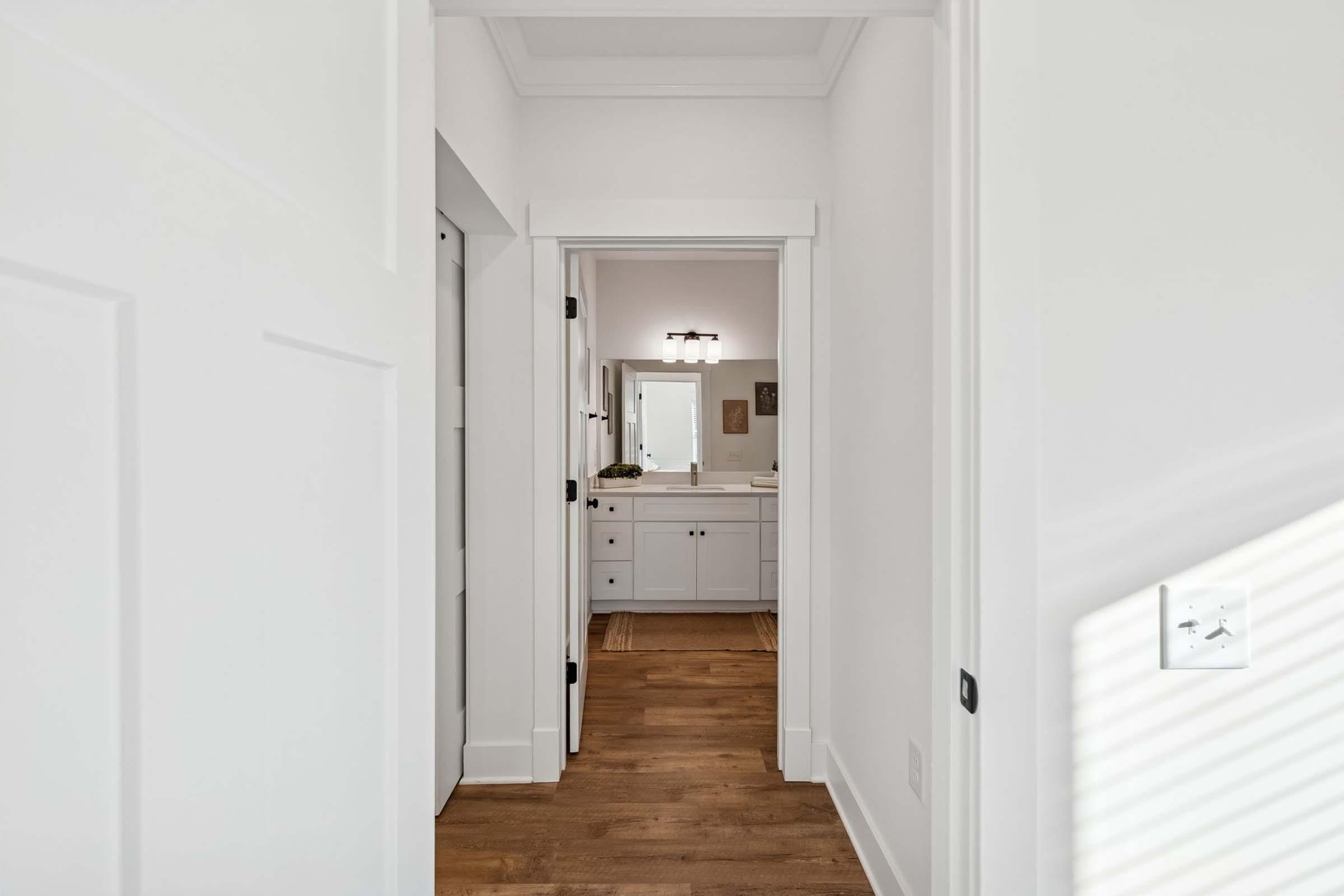 A bright, modern hallway leading to a bathroom. The walls are painted white, and the wooden floor adds warmth. In the bathroom, a mirror and double sink can be seen along with minimal decorative elements. Natural light filters through a nearby window, enhancing the airy feel of the space.