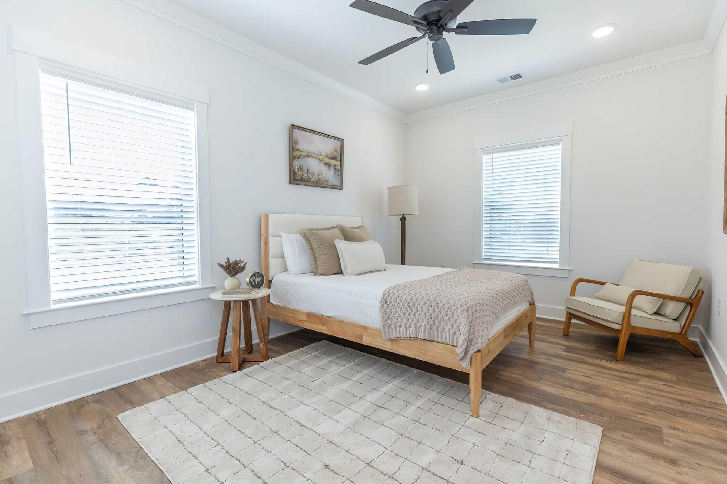 A modern bedroom featuring a wooden bed with a light-colored blanket, a round side table with a plant, an armchair, and a lamp. The room has two windows with white blinds, a light wooden floor, and a neutral color palette, creating a spacious and airy atmosphere. A painting hangs on the wall above the bed.