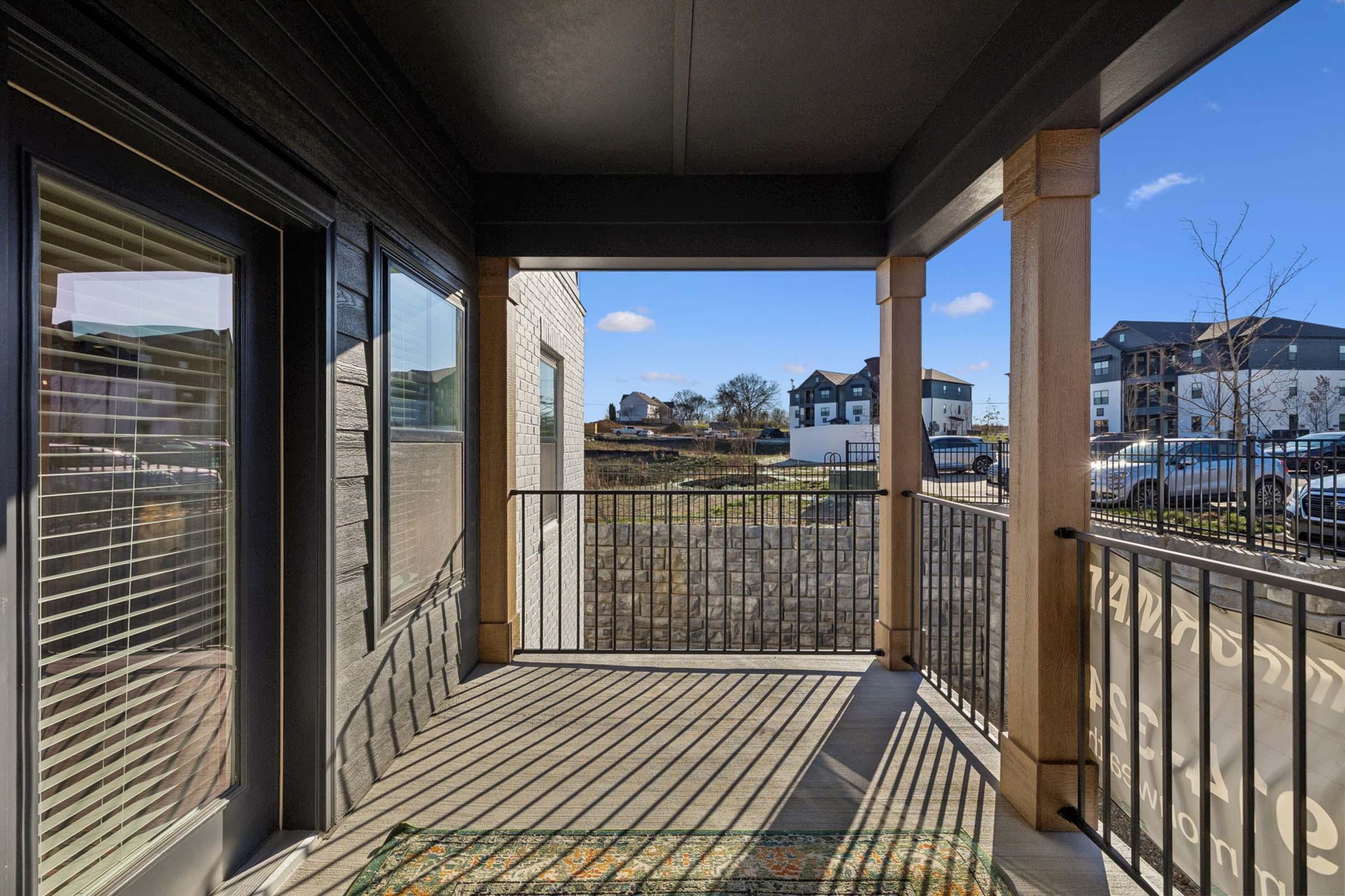 A view from a covered balcony looking out onto a residential area. The balcony features wooden posts and a metal railing, with sunlight creating shadows on the flooring. In the distance, there are apartment buildings and parked cars. The sky is clear with a few clouds.