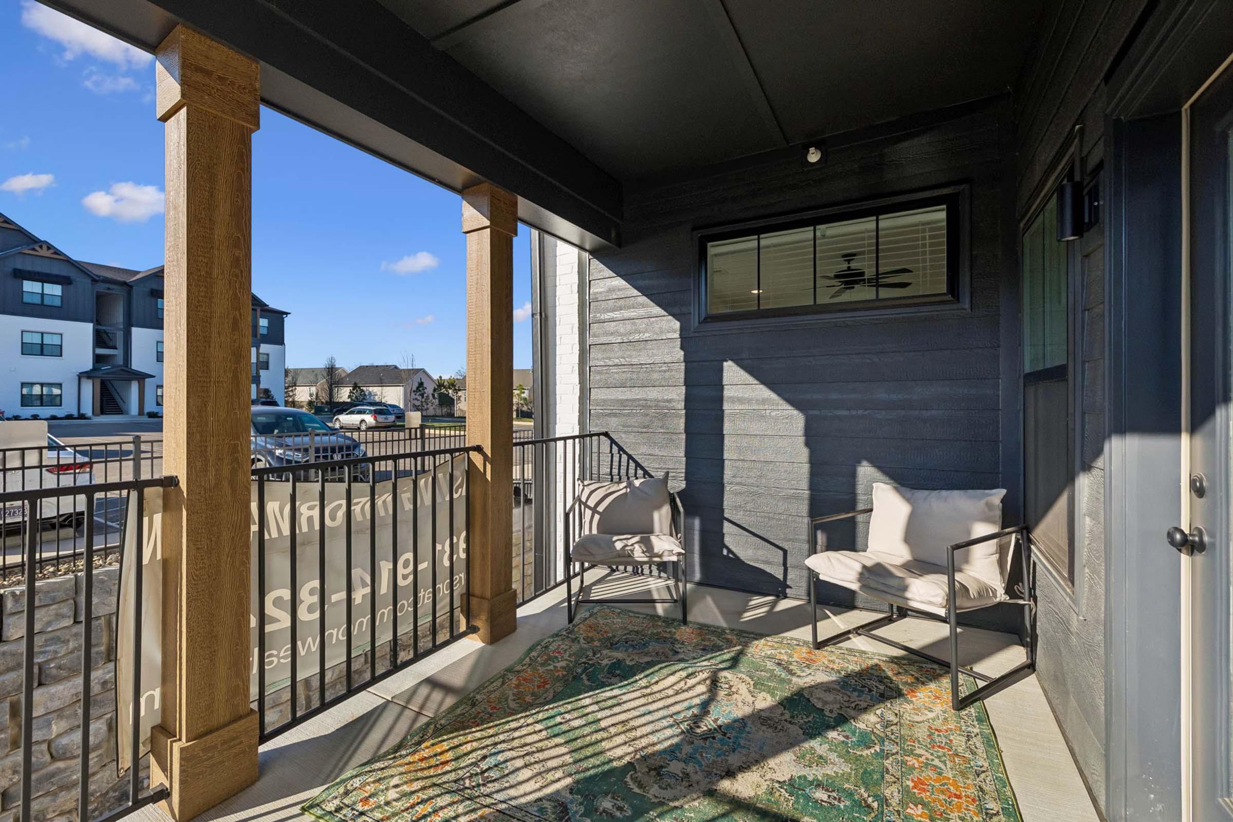 A covered outdoor patio with two chairs and a colorful rug, featuring a view of nearby buildings and a clear blue sky. The scene conveys a welcoming, cozy atmosphere.