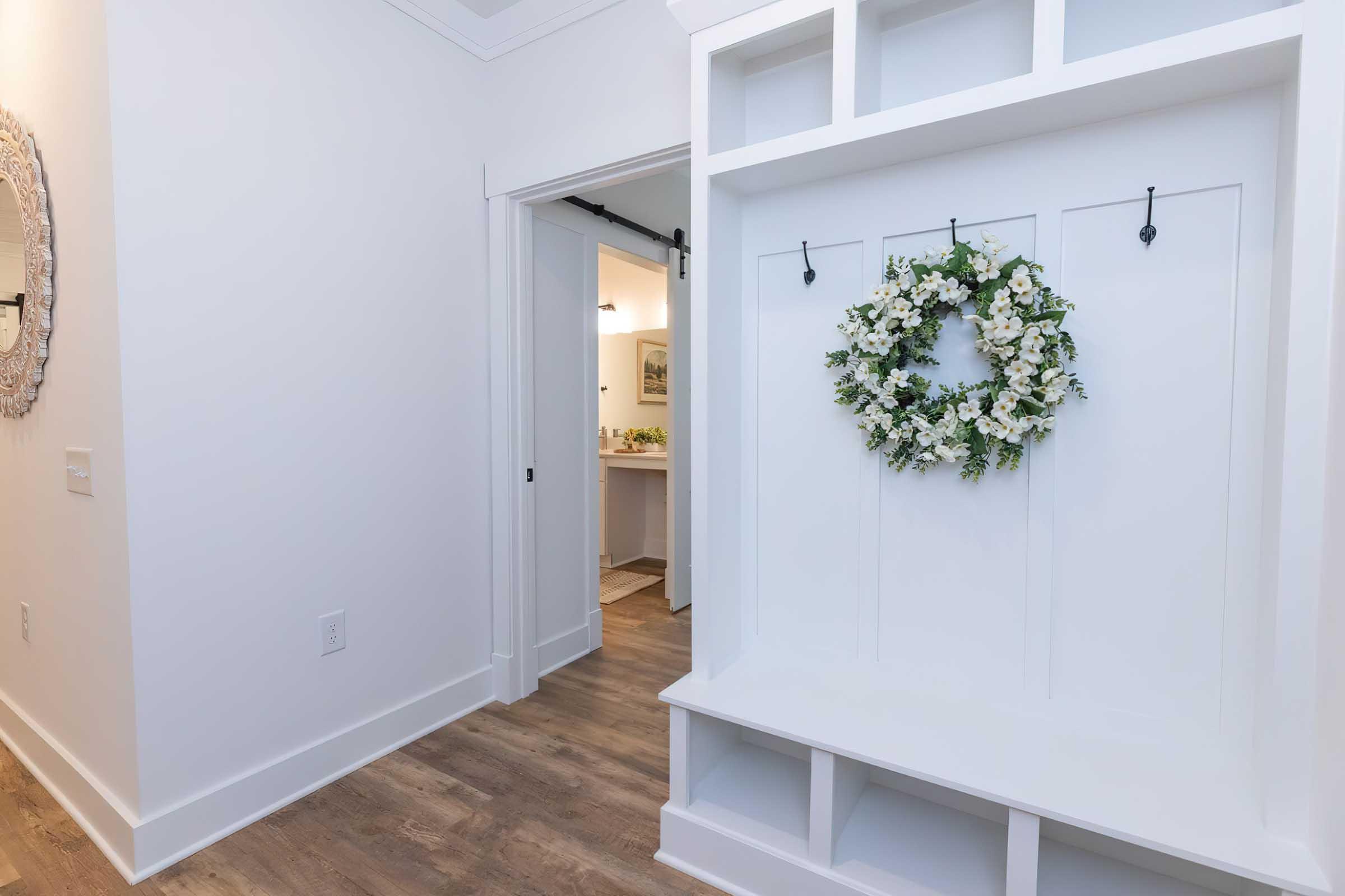A bright and modern entryway featuring a built-in bench with hooks. A decorative wreath made of white flowers hangs on the wall. The walls are painted white, and the floor has light wooden planks. A doorway leads to another room, partially visible in the background.
