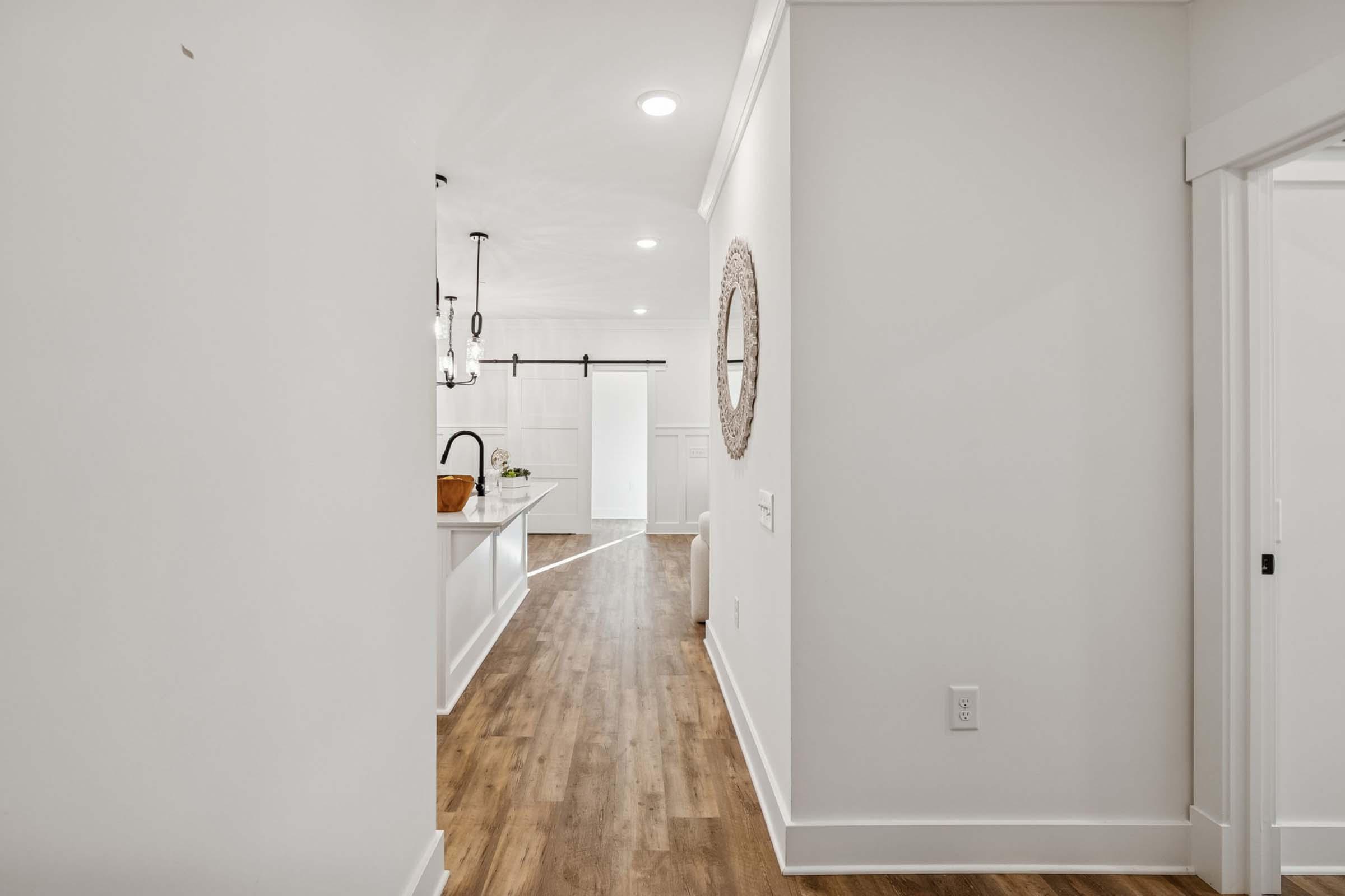 A bright, modern hallway featuring light-colored walls and hardwood flooring. A round mirror hangs on one wall, while the other side showcases a glimpse of a kitchen with white cabinetry and stylish lighting fixtures. The space is well-lit with recessed lights, creating an airy and inviting atmosphere.