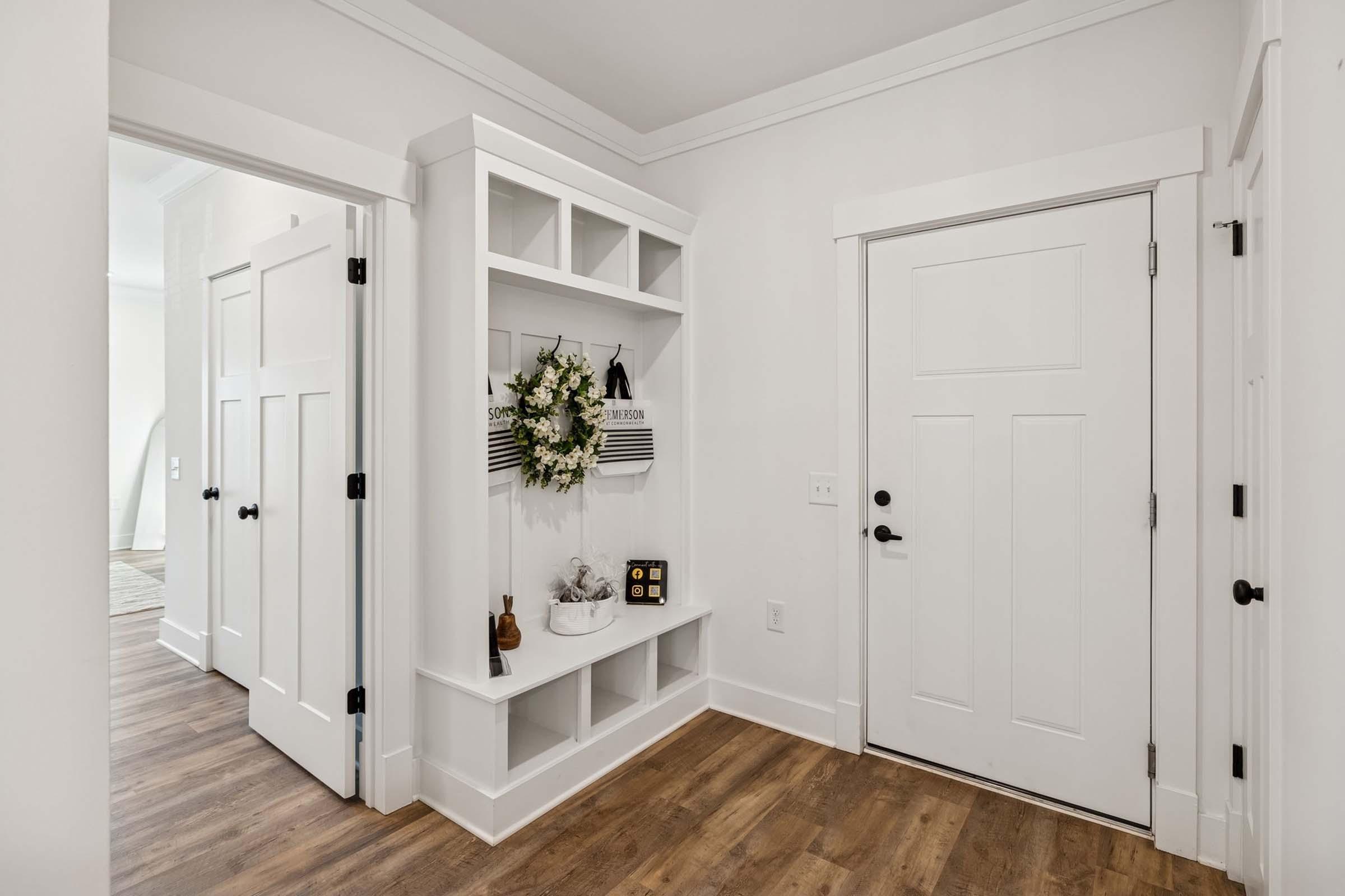 A bright and airy entryway featuring a white door and a shelving unit against the wall. The shelving unit displays decorative items and a wreath. Light wood flooring complements the modern design. Two doors lead to adjacent rooms, enhancing the open feel of the space.