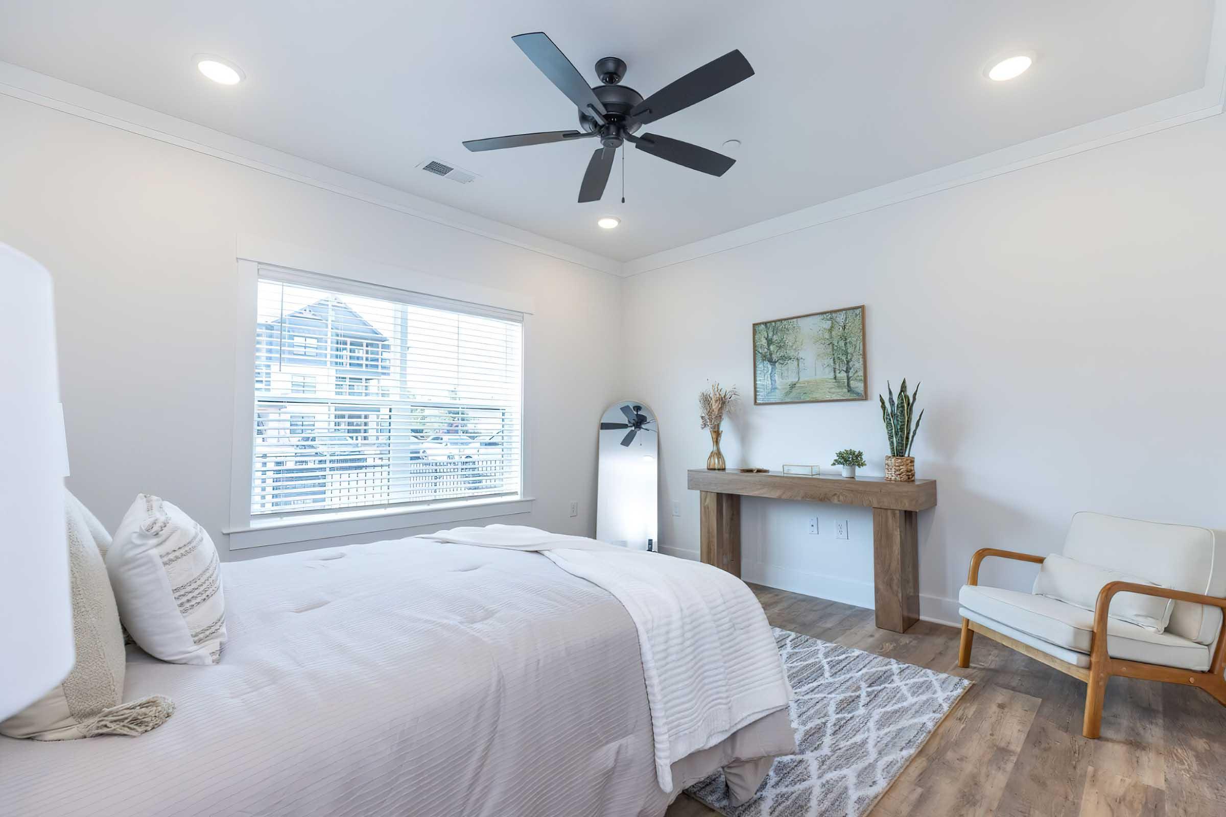 A modern, well-lit bedroom featuring a queen-sized bed with light bedding, a ceiling fan, and a large window with blinds. There's a wooden table adorned with decorative plants and a piece of artwork on the wall. A cozy armchair is placed beside the table, adding to the room's inviting atmosphere.