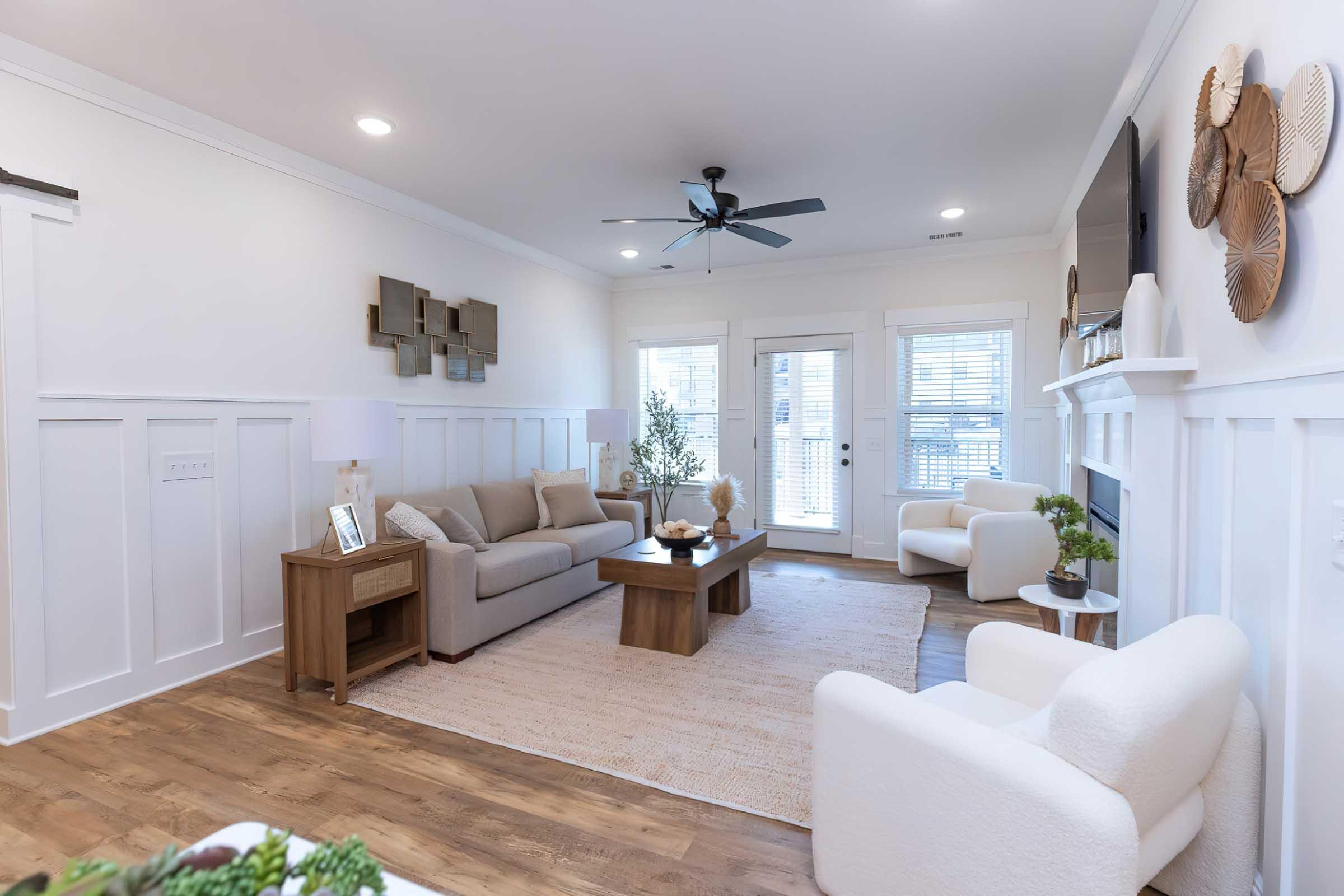 A bright and modern living room featuring a light beige sofa, a wooden coffee table, and two plush white chairs. The room has white wainscoting, a light wood floor, and a ceiling fan. Natural light streams in through large windows, and there are decorative elements like wall art and plants.
