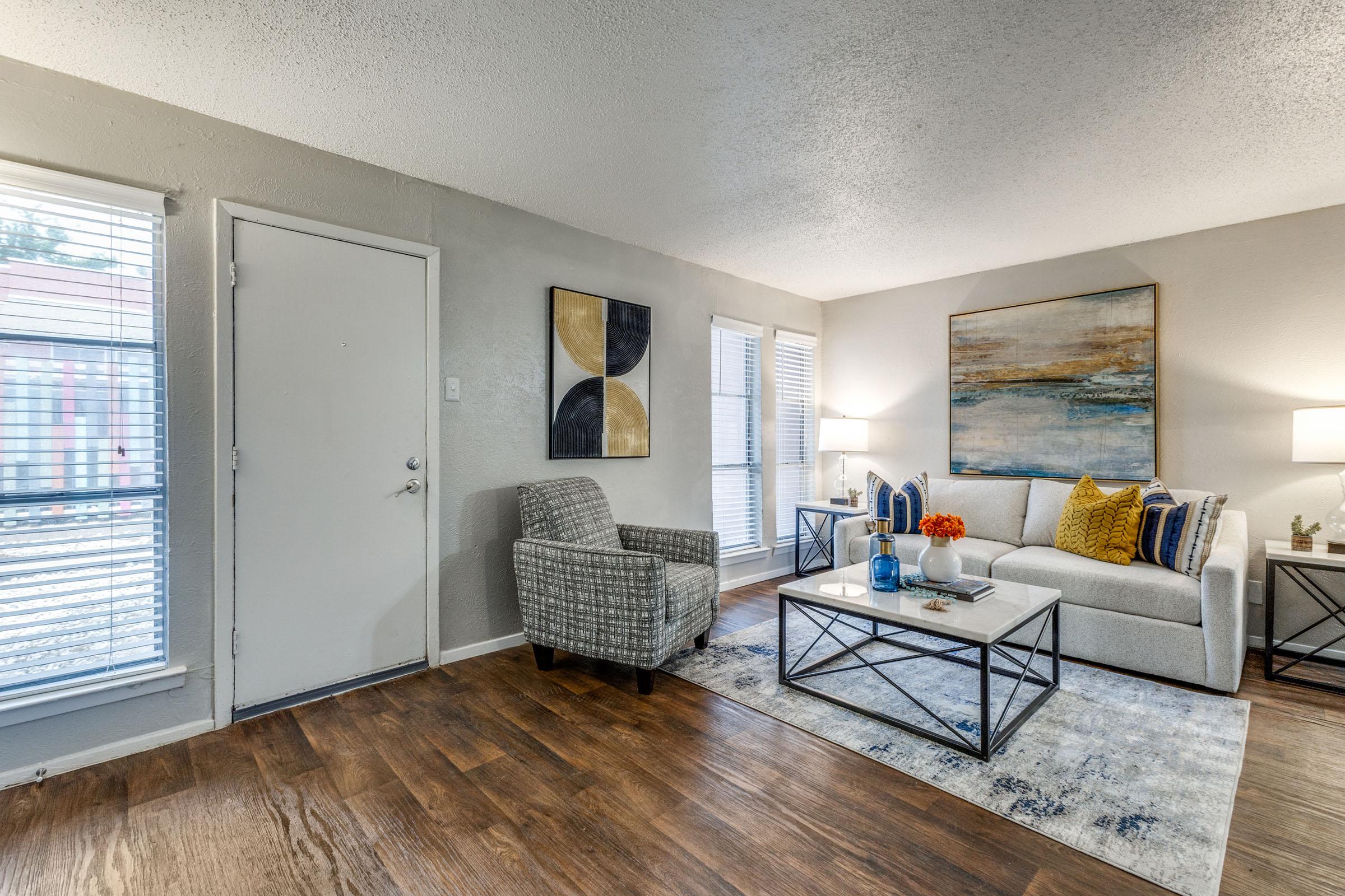 A modern living room featuring a gray sofa with decorative pillows, a coffee table with a blue vase and orange flowers, and a comfortable armchair. The decor includes abstract artwork on the wall and large windows with blinds, allowing natural light to fill the space. The floor is wooden.