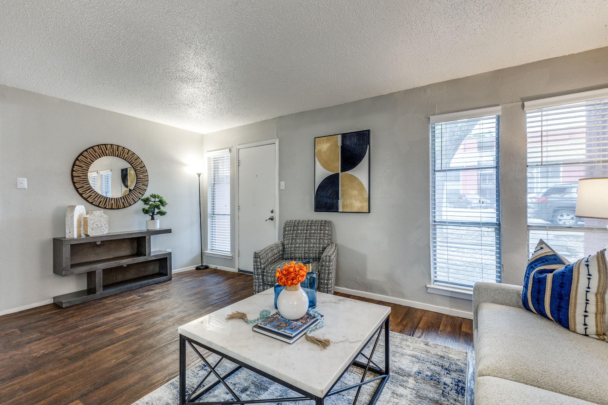 A cozy living room featuring a light grey wall, wooden flooring, and large windows. The space includes a modern grey sofa with blue and white cushions, a marble coffee table with decorative items, a stylish circular mirror, and abstract wall art. Natural light enhances the inviting atmosphere.