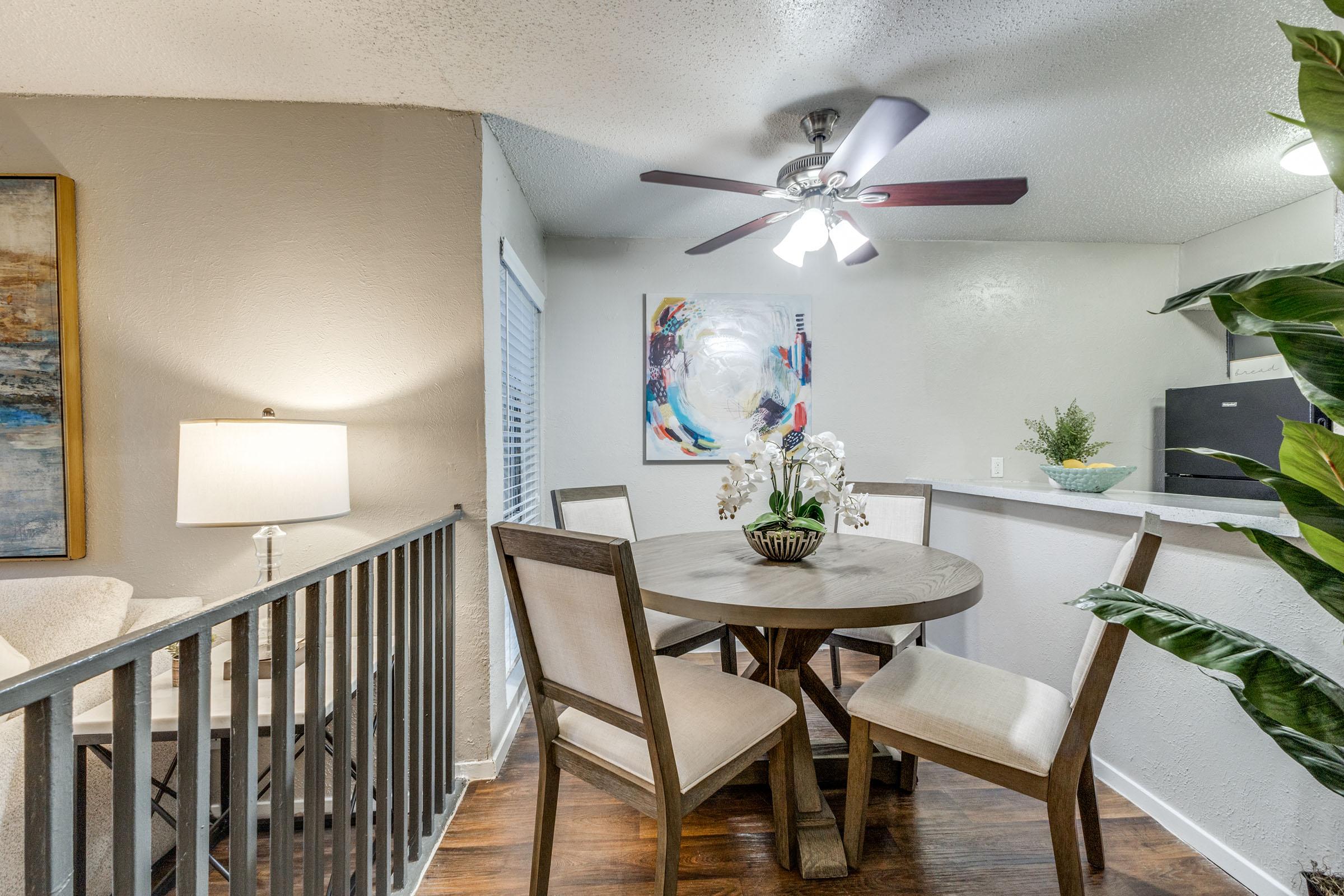 A well-lit dining area features a round wooden table with four upholstered chairs, a potted orchid centerpiece, and a ceiling fan. In the background, there’s a colorful abstract painting, a window with blinds, and a small kitchen area visible. A soft lamp adds warmth to the space.