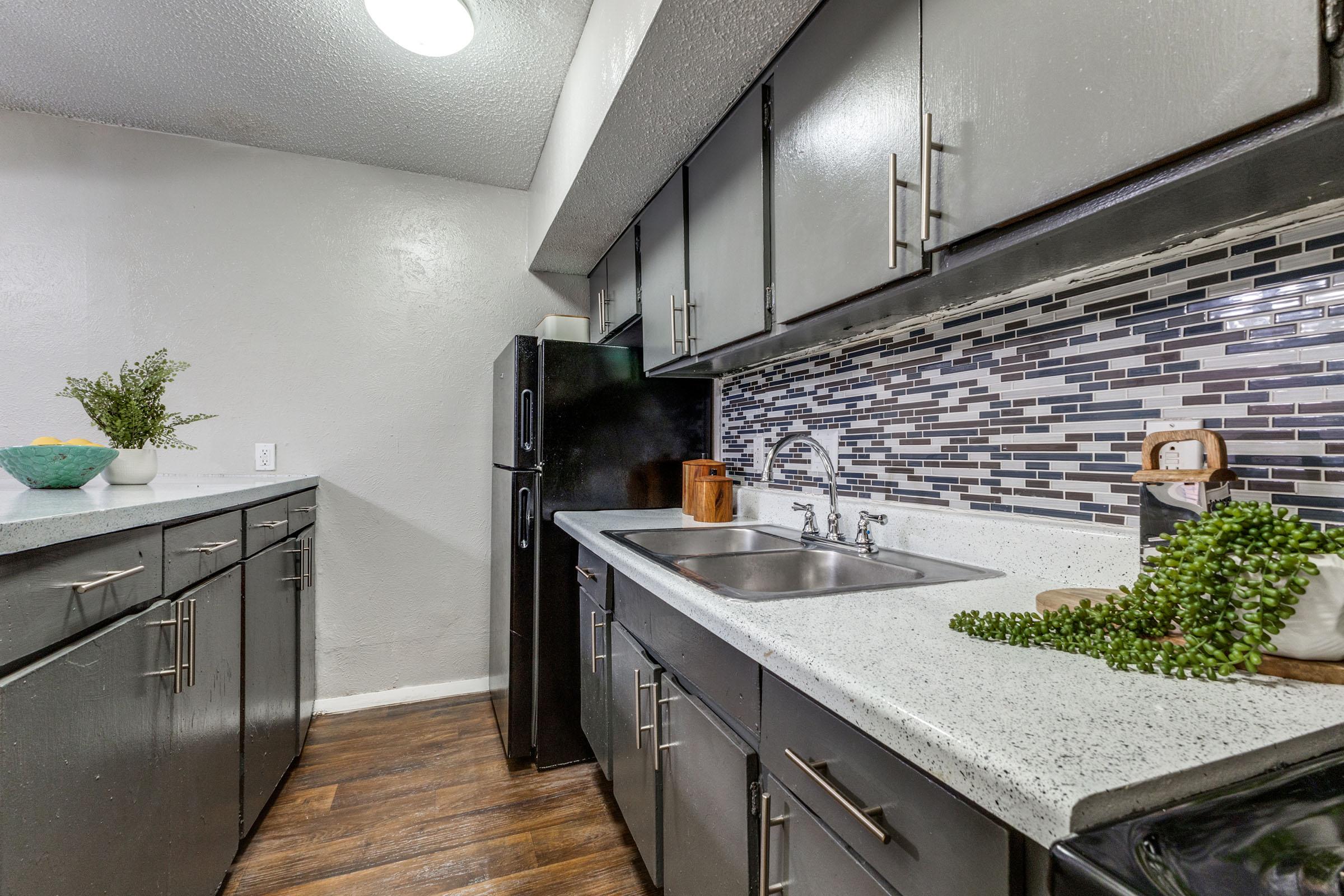Modern kitchen featuring gray cabinets, a black refrigerator, a double sink, and a decorative backsplash made of mosaic tiles. Countertops made of a light-colored stone with a small plant and wooden kitchen accessories arranged on it. The flooring is wood-like laminate.