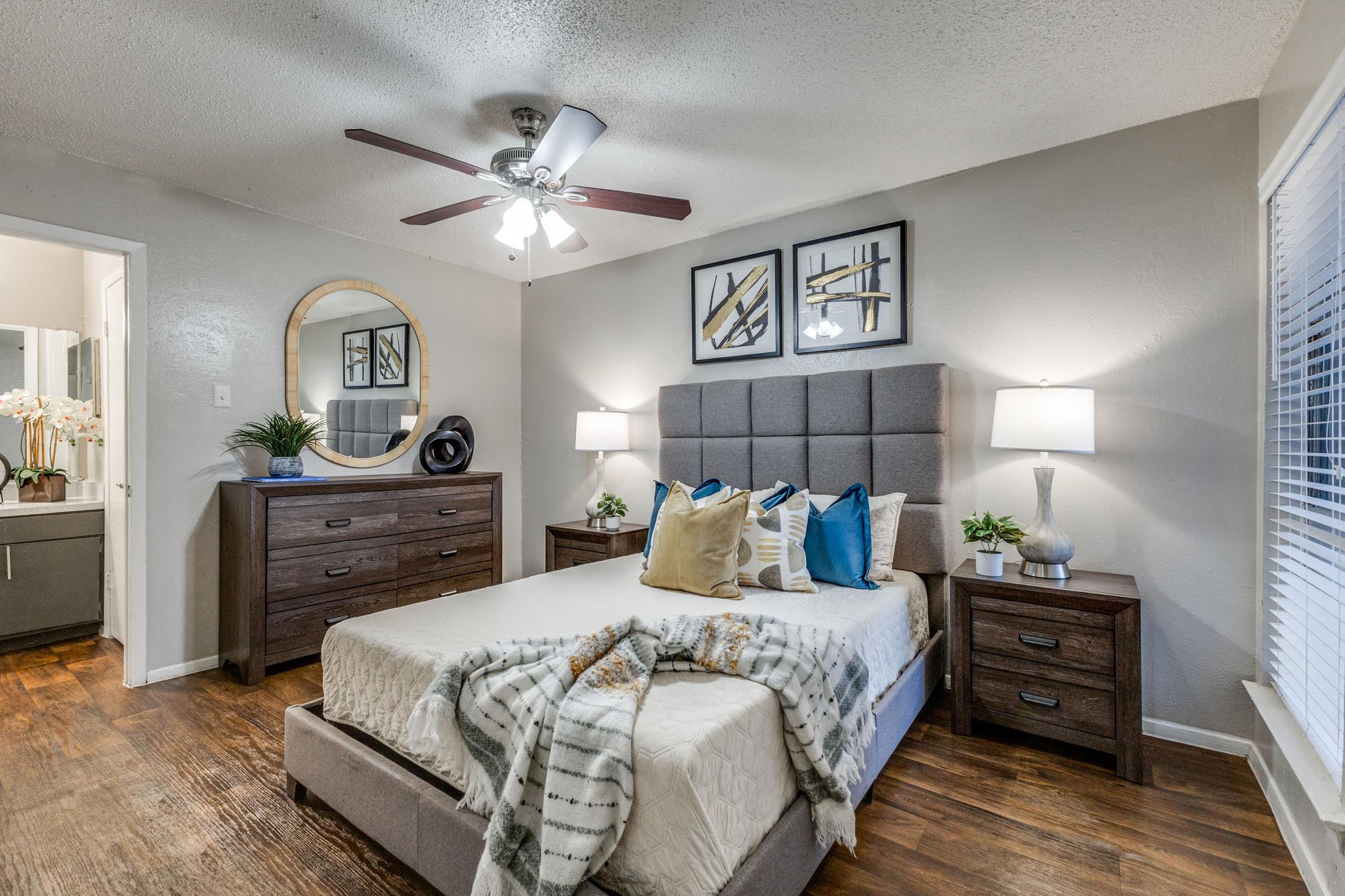 A modern bedroom featuring a gray upholstered bed with decorative pillows, a cozy blanket, and wooden furniture including a dresser and nightstands. Two lamps provide soft lighting, while a circular mirror hangs on the wall. A ceiling fan adds to the contemporary decor, and there are stylish art pieces on the walls.