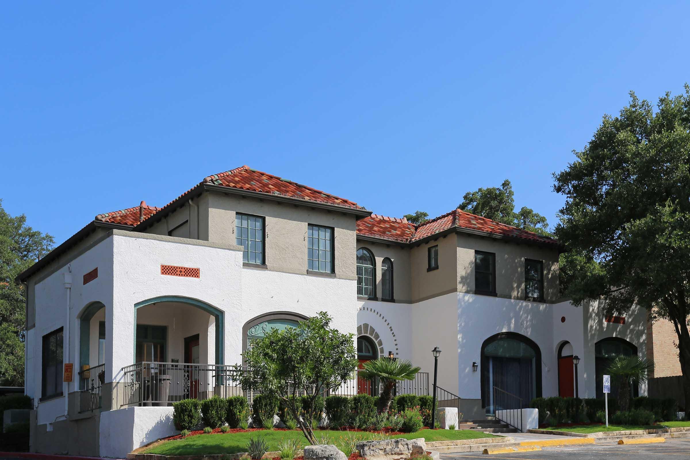 A two-story building with a Mediterranean architectural style features a white exterior with decorative elements, red-tiled roofs, and large windows. The structure is surrounded by greenery, including shrubs and trees, against a clear blue sky.