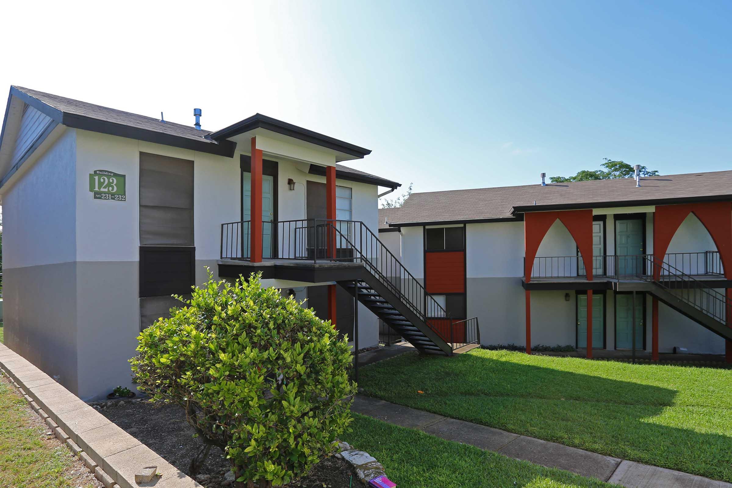 Two apartment buildings side by side in a residential area. The building on the left features a white and red exterior with a staircase, while the building on the right has a similar architectural style with large windows and a manicured lawn in front. Bright blue sky in the background.