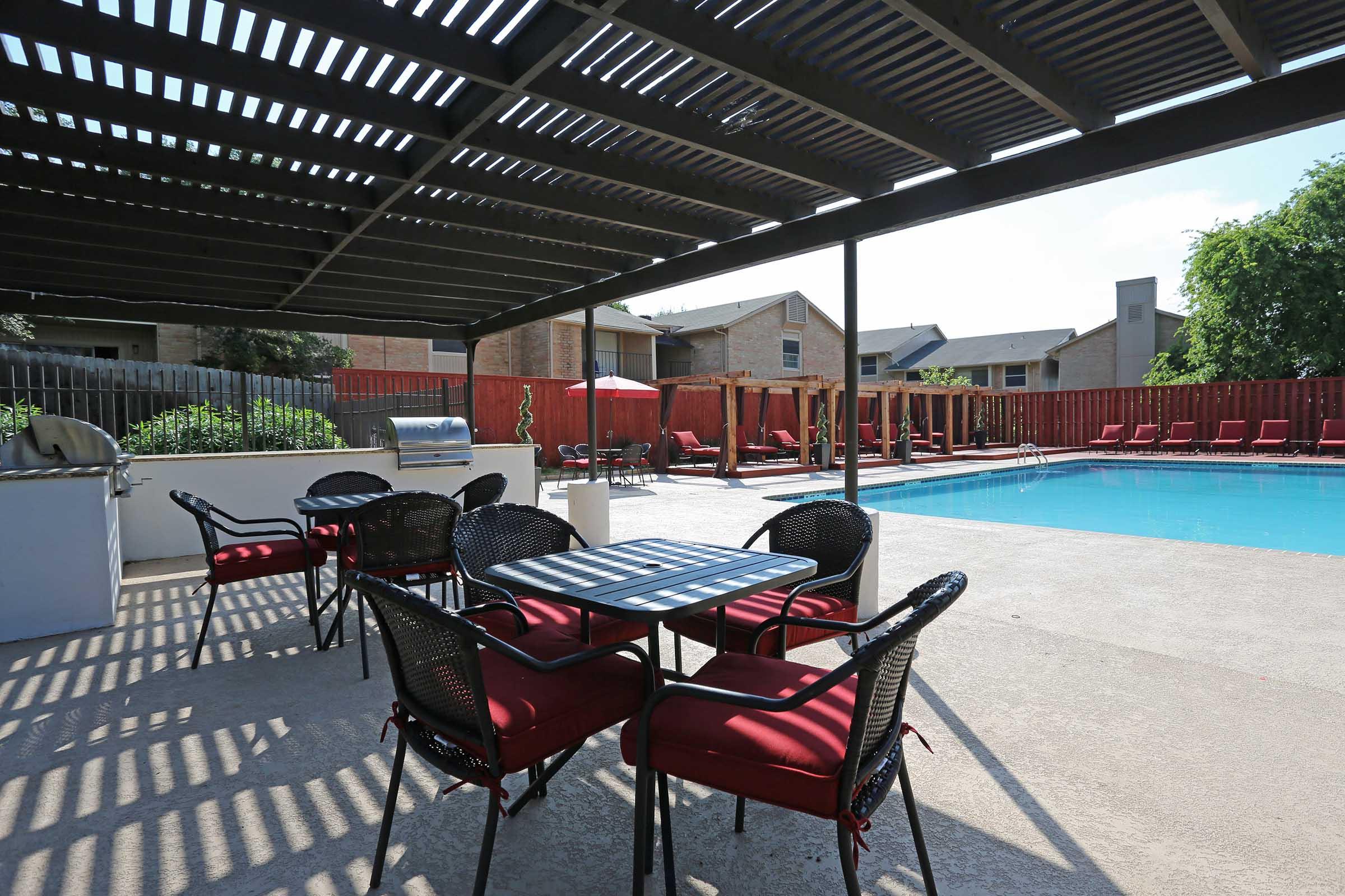 An outdoor pool area featuring a blue swimming pool, surrounded by a concrete deck. There are several black chairs with red cushions and a table in the shade of a pergola. In the background, there are residential buildings and a wooden fence, creating a cozy atmosphere for relaxation and leisure.