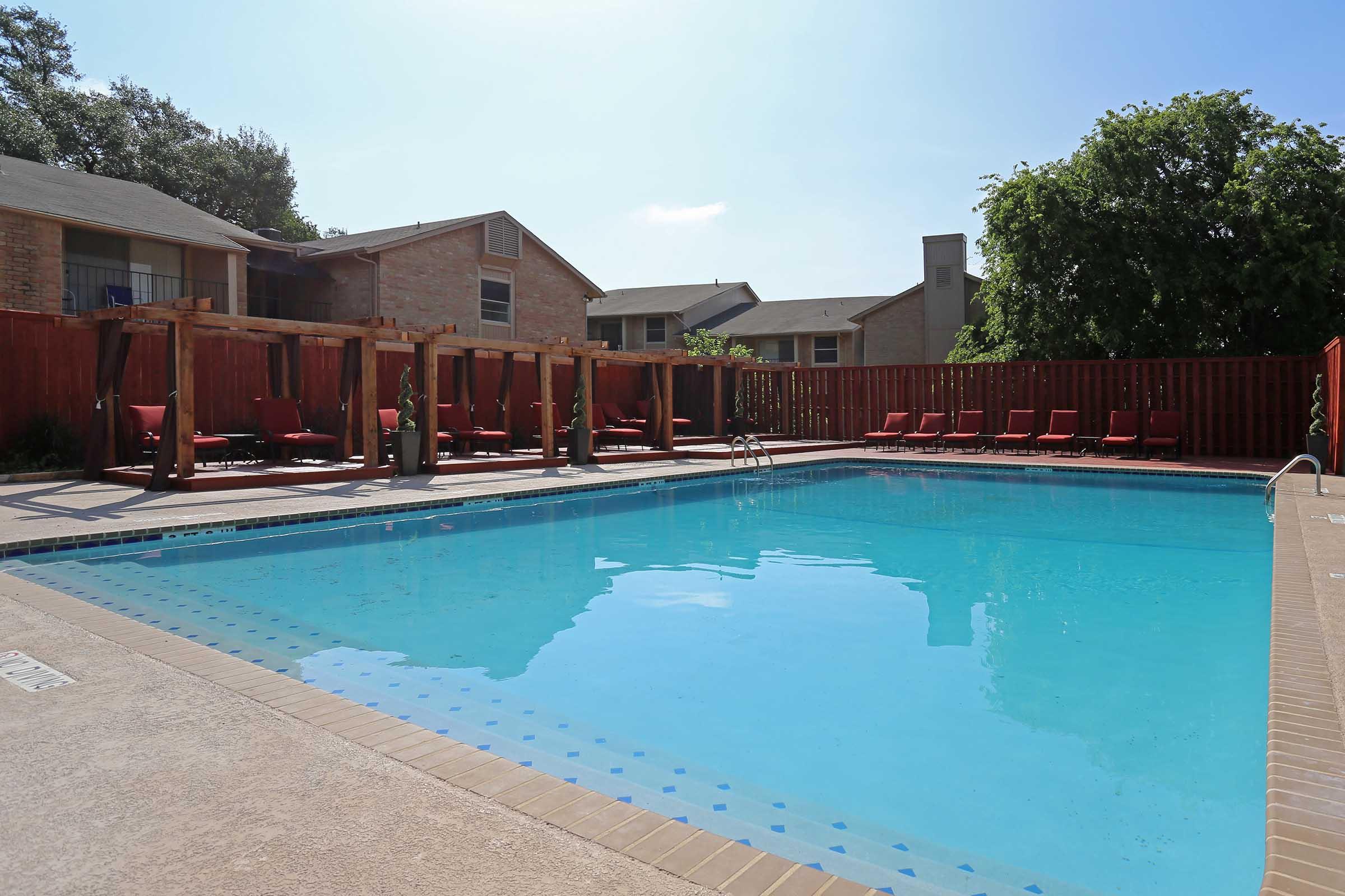 A clear, blue swimming pool surrounded by lounge chairs on a sunlit day. Wooden pergolas provide shade along one side, with a brown privacy fence in the background. The scene is set in a residential area with buildings visible nearby. The atmosphere is inviting and relaxing.