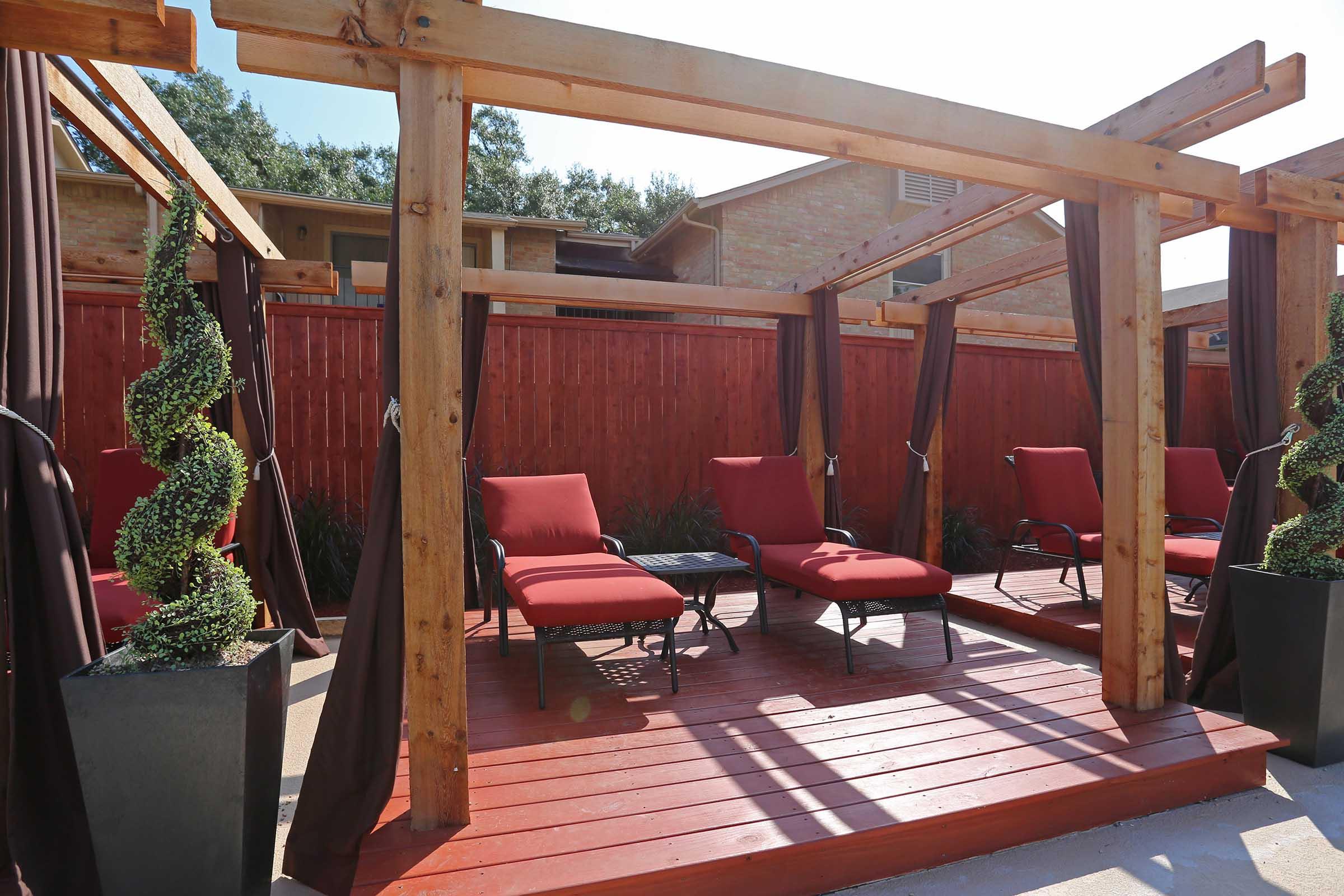 A cozy outdoor seating area featuring red lounge chairs under wooden pergolas. The space is decorated with potted spiral topiary plants and has a wooden deck. Sunlight filters through, creating a warm and inviting atmosphere. A red wooden fence provides privacy in the background.