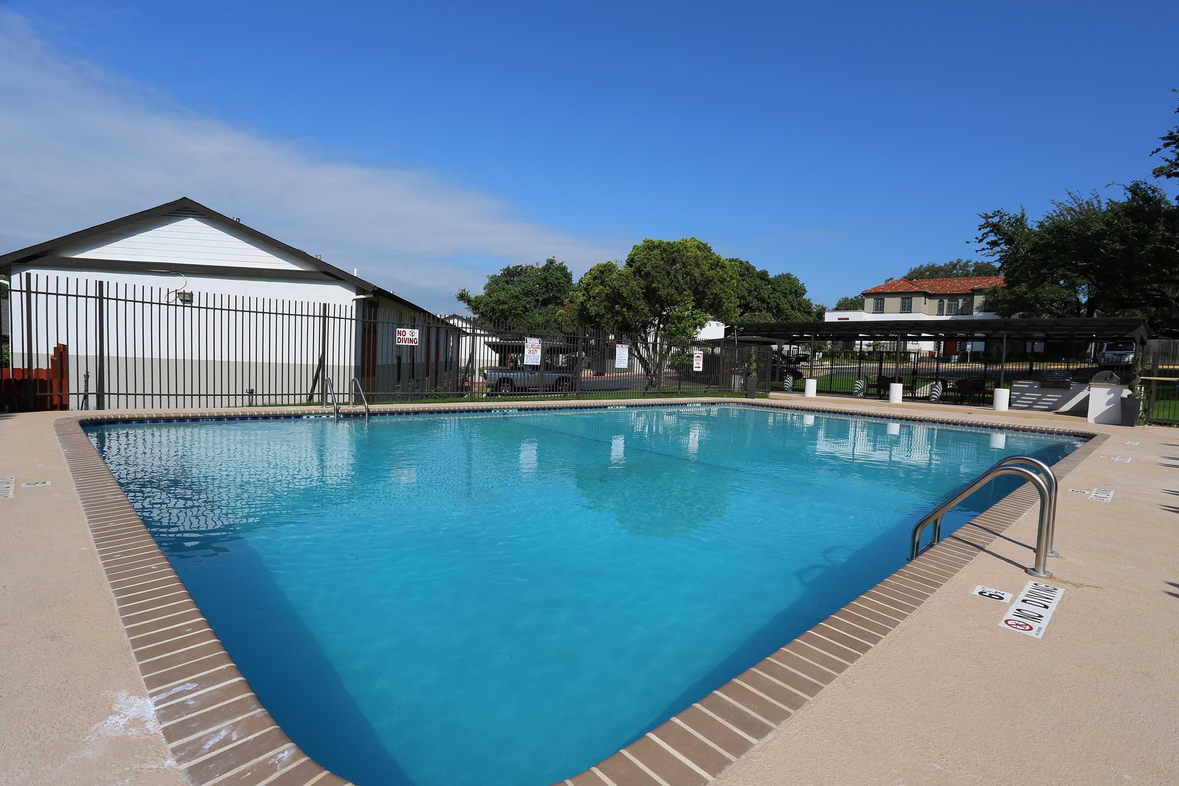 A clear blue swimming pool surrounded by a tan deck. There are shaded seating areas with picnic tables and a building in the background. The sky is partly cloudy, creating a bright and inviting atmosphere. A fence encloses the pool area for safety.