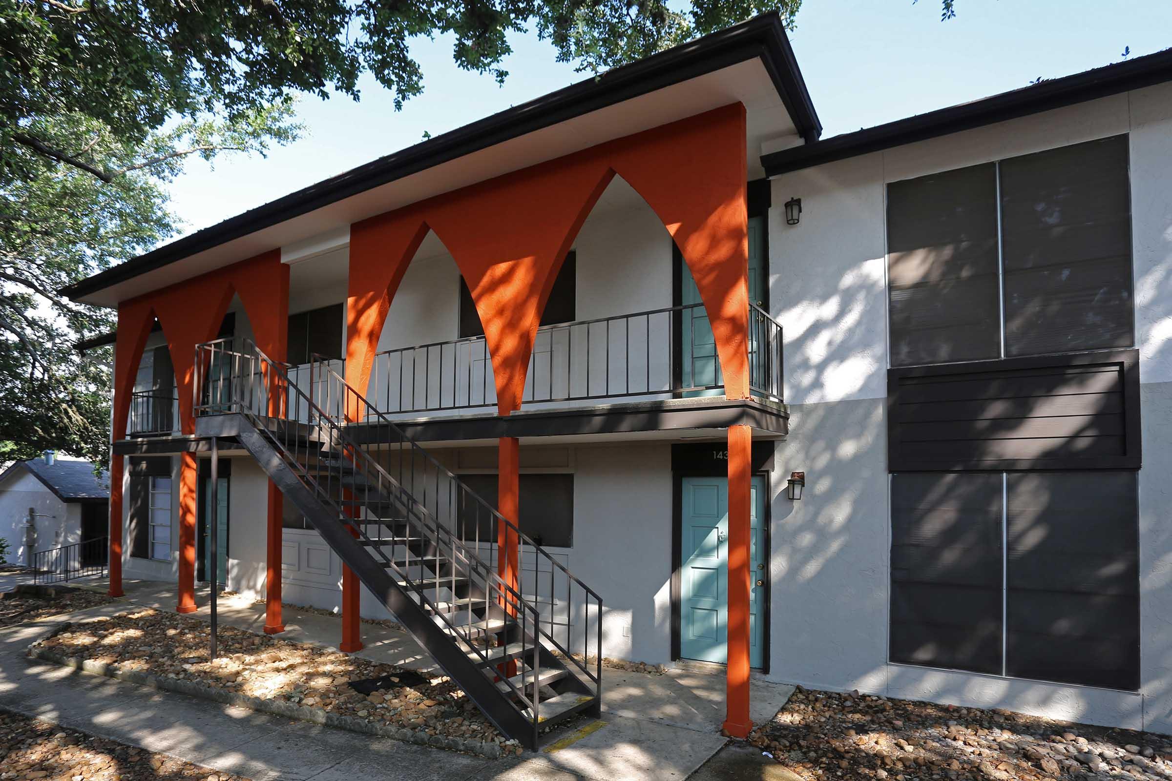 Two-story apartment building featuring a modern architectural design with bold orange arches and a staircase leading to the second floor. The ground level has minimalist doors, and the walls are a blend of white and grey tones. Surrounding greenery enhances the contemporary aesthetic.