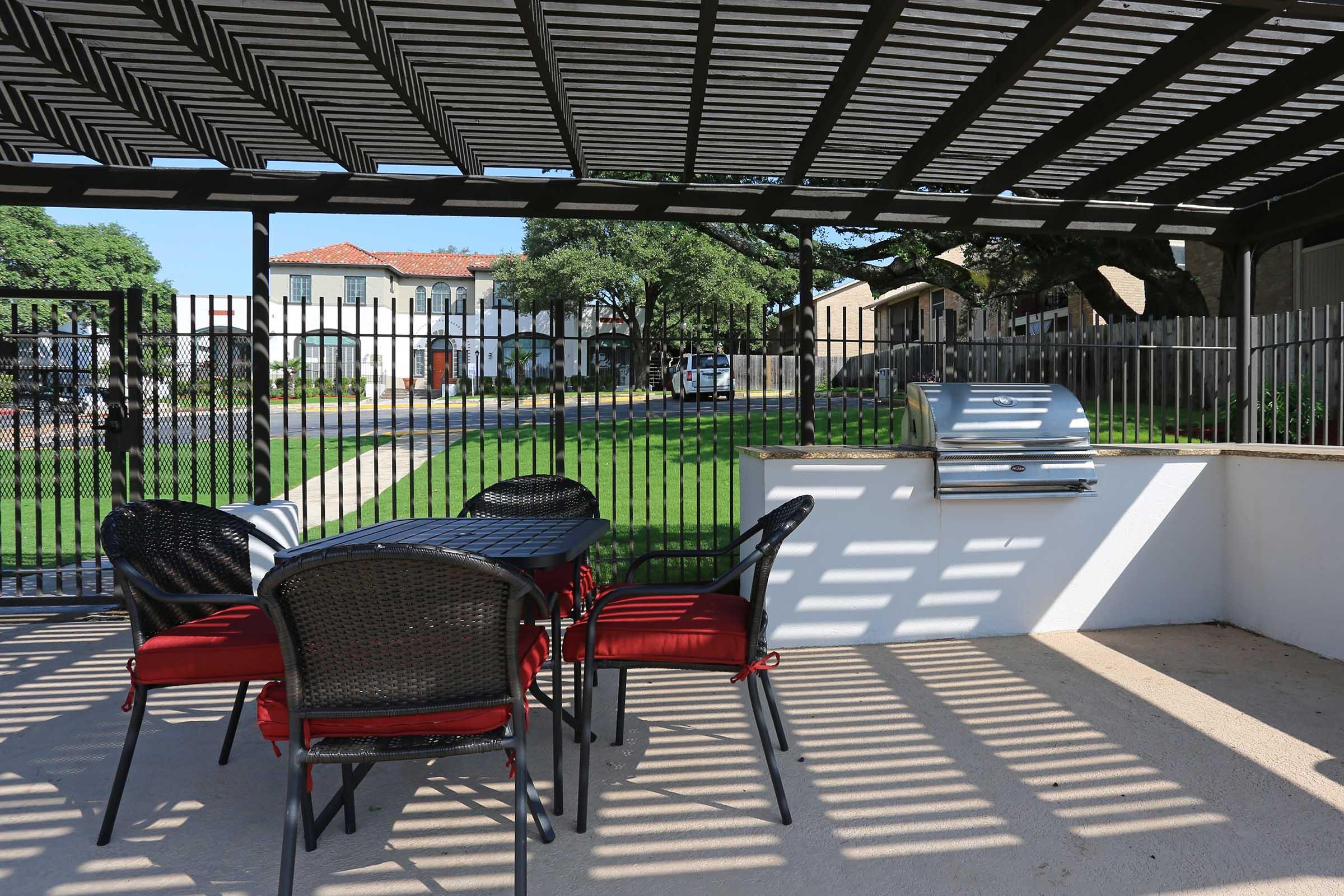Outdoor patio area with a shaded seating arrangement featuring a table and four chairs, alongside a built-in grill. The background includes a grassy area and a decorative fence. Sunlight creates a pattern through the overhead slats, enhancing the inviting atmosphere of the space.
