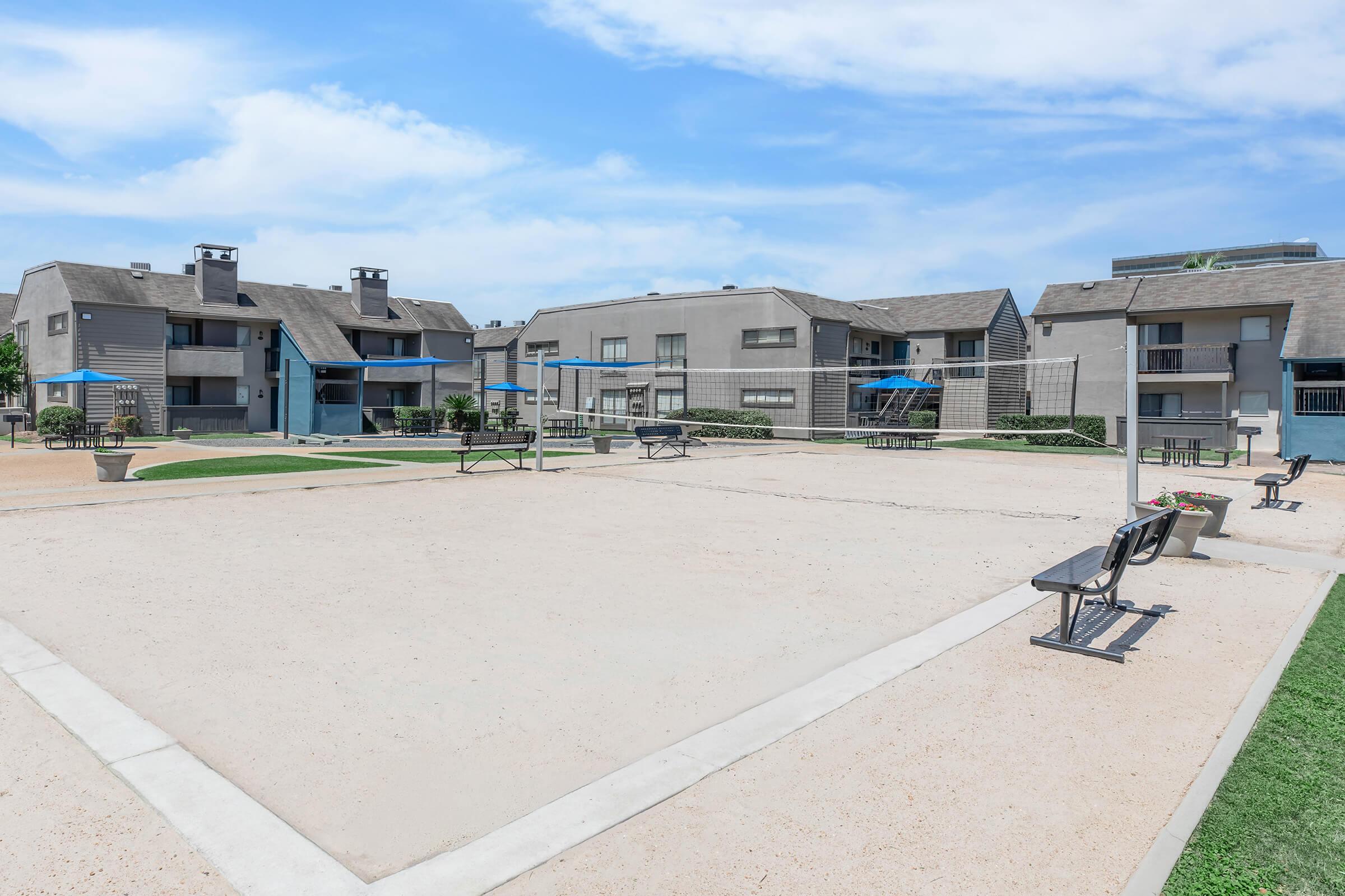 A sandy volleyball court in an apartment complex, surrounded by grey multi-unit buildings. There are benches along the court and some umbrellas for shade. The sky is clear with a few clouds, and the area is well-maintained with green grass.