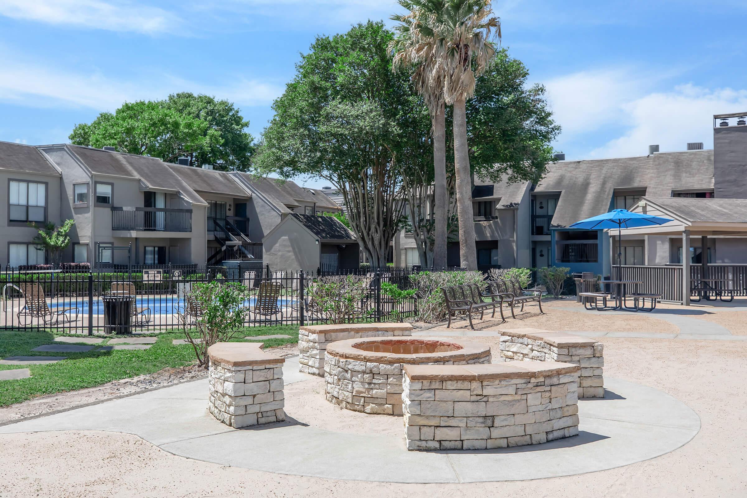 A landscaped area featuring a circular stone fire pit surrounded by benches, with palm trees in the background. Nearby, there are apartment buildings with balconies, a swimming pool, and outdoor seating under a blue umbrella, all set in a bright, sunny environment.