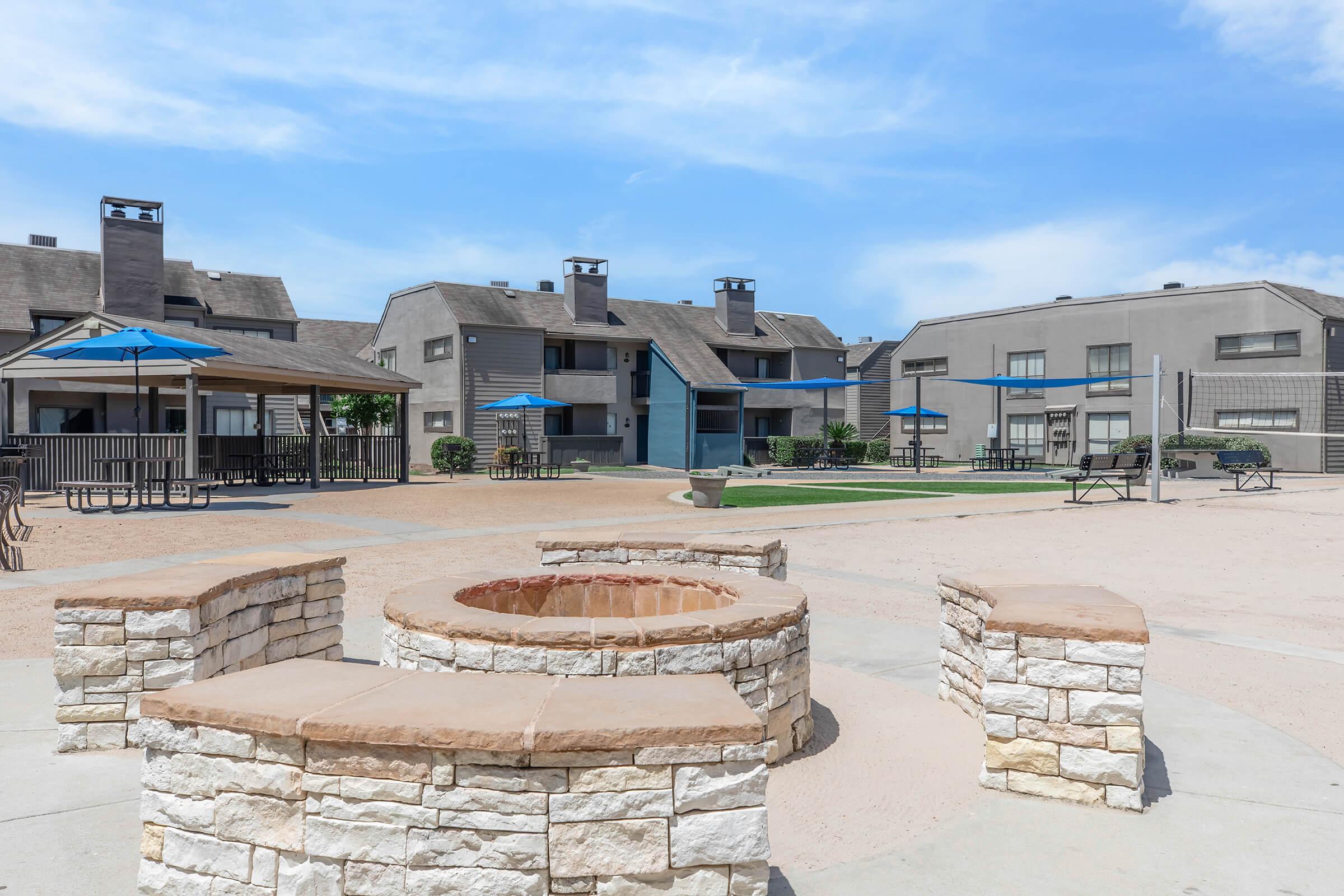 A view of a residential community featuring several grey apartment buildings, outdoor seating areas with blue umbrellas, and a stone fire pit. The surrounding grounds are sandy with grass patches, under a bright blue sky with scattered clouds.