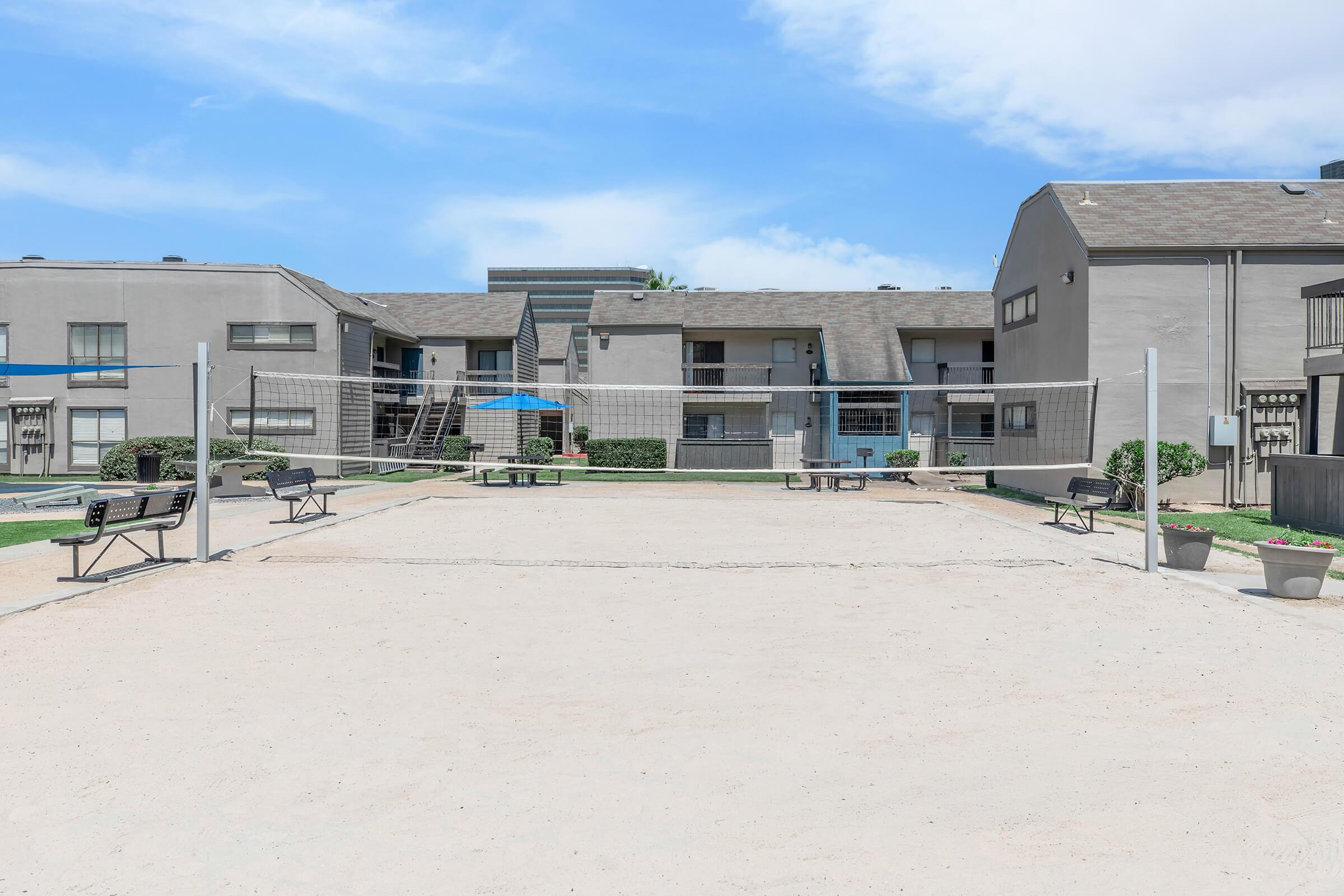 A sandy volleyball court surrounded by low-rise apartment buildings. Benches are placed on either side of the court, and there are potted plants in the foreground. The sky is clear with a few clouds, creating a bright and inviting atmosphere.