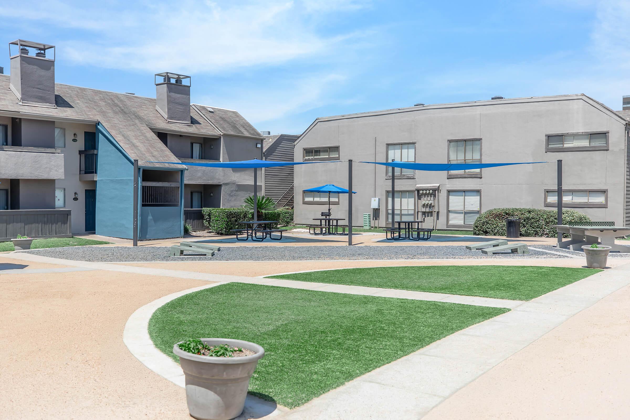 A view of a well-maintained outdoor courtyard at an apartment complex featuring shaded seating areas with blue umbrellas, surrounded by gravel and green grass patches. Modern buildings in the background complete the inviting atmosphere, ideal for relaxation and socializing.