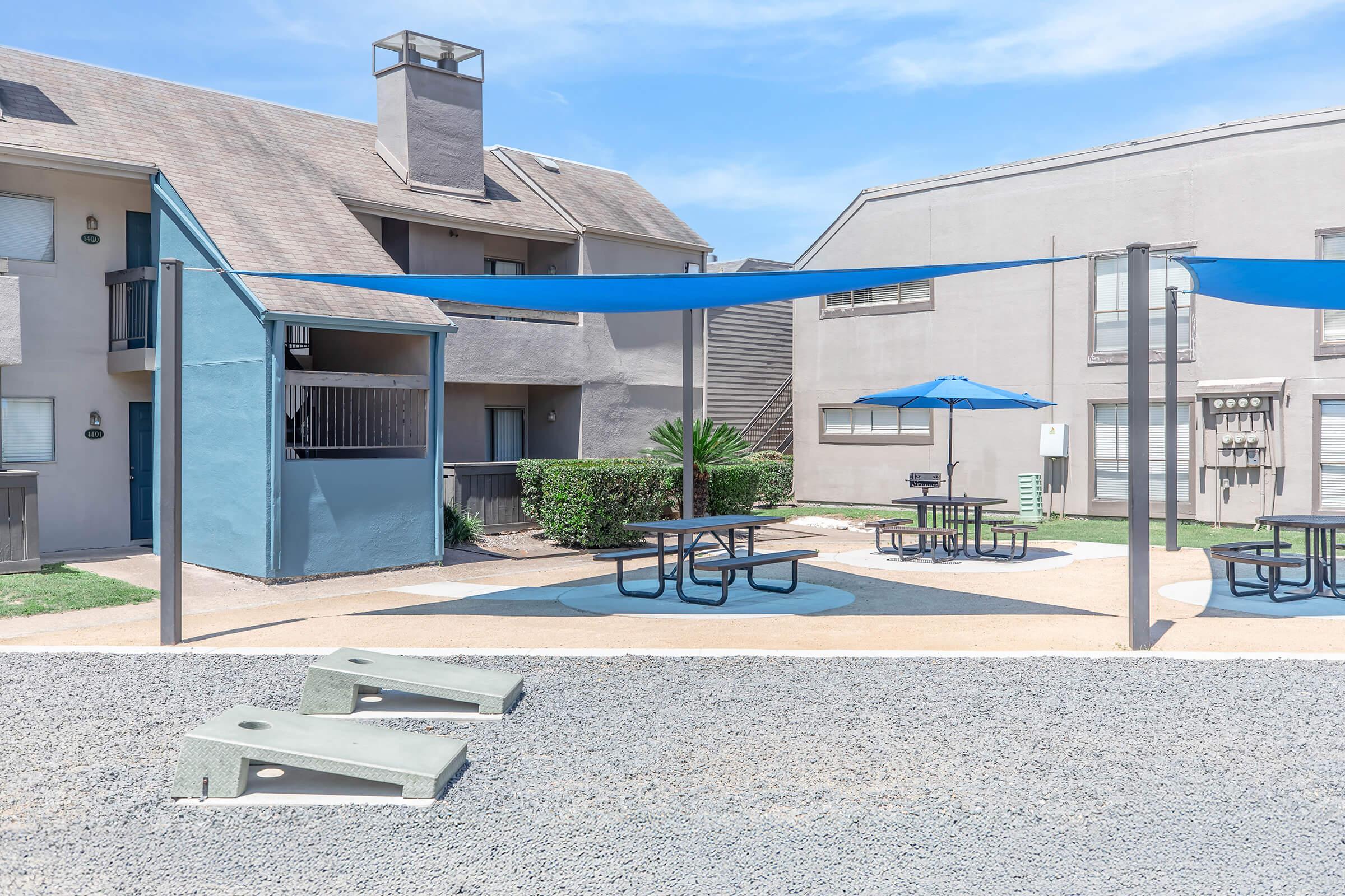 A landscaped outdoor area featuring picnic tables under blue shade sails, a blue umbrella table, and a grill. Nearby, there are two concrete seating structures in a gravel setting, alongside multi-story residential buildings and green shrubbery. Clear blue skies enhance the scene.